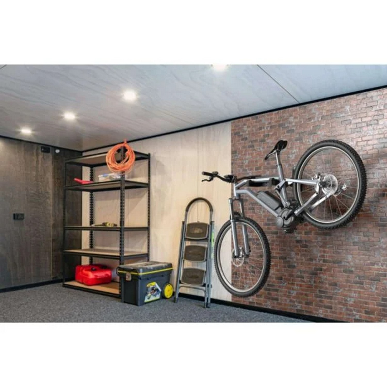 Un vélo est monté sur un mur de briques. Une étagère métallique avec des outils et un récipient rouge. Une échelle et une boîte à outils.