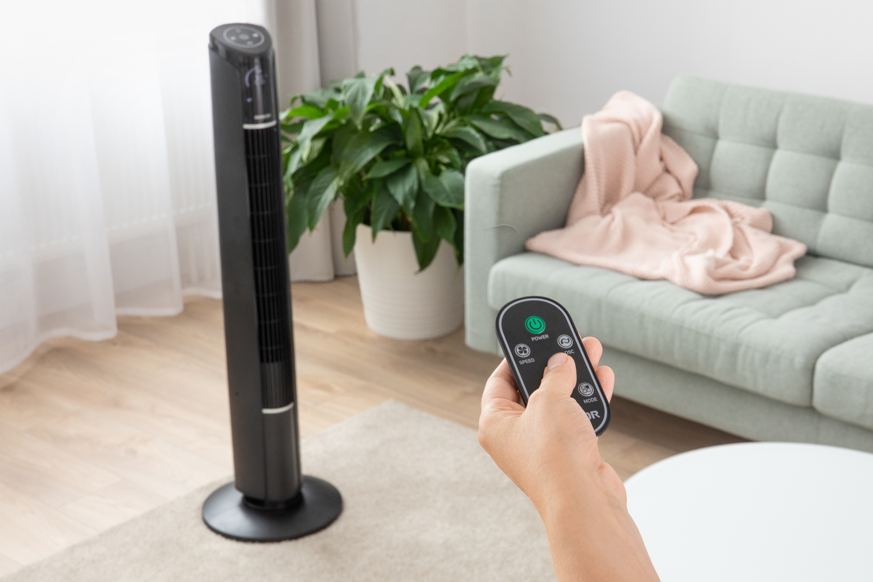 Fekete toronyventilátor távirányítóval egy szobában, kanapéval és növénnyel.