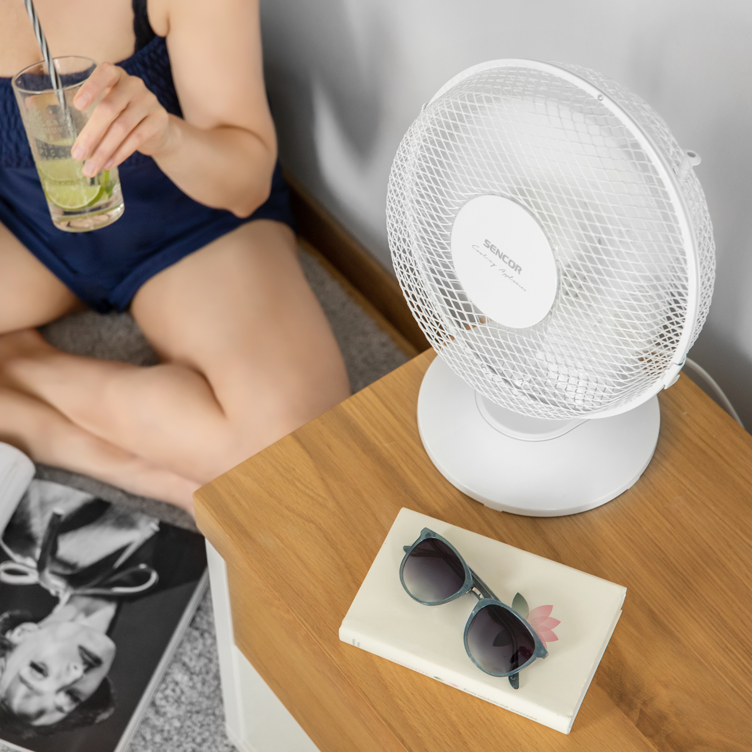 Egy nő itallal ül egy fehér ventilátor mellett egy faasztalon. Napszemüveg és könyv a közelben.