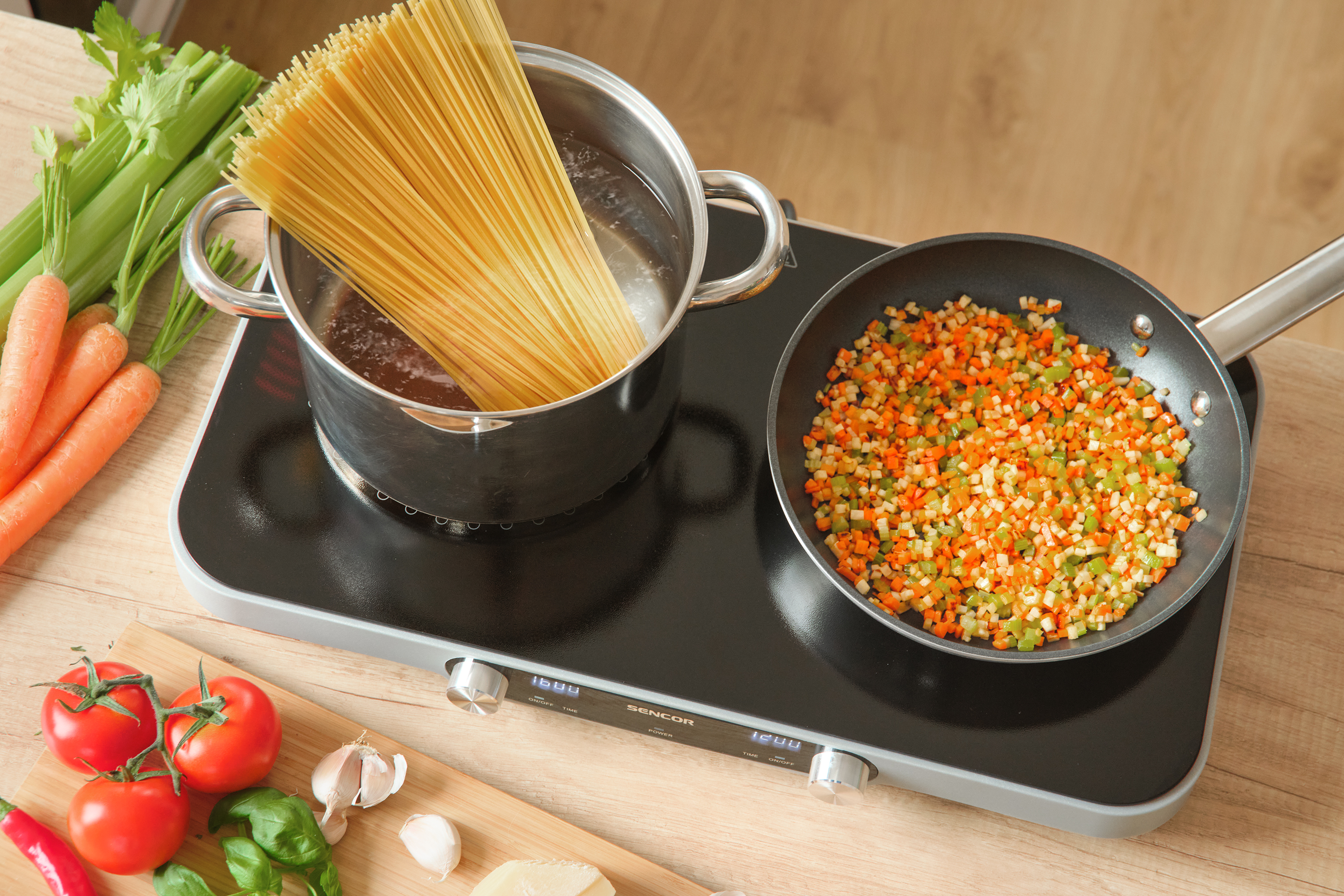 Spagetti a fazékban, zöldségek a serpenyőben, fekete főzőlapon. Paradicsom, fokhagyma, bazsalikom és egyéb hozzávalók.