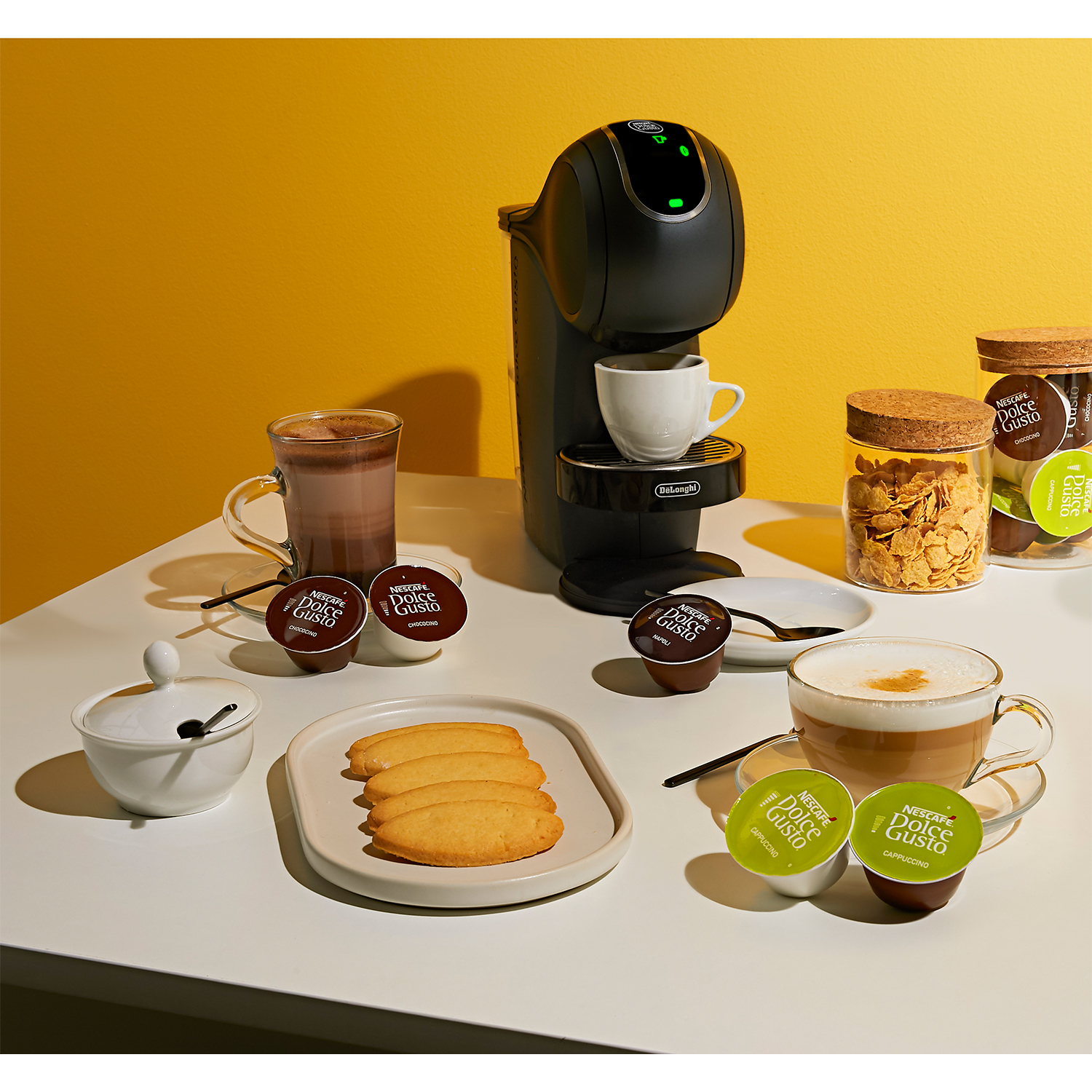 Macchina da caffè con tazze, biscotti e capsule su un tavolo bianco.