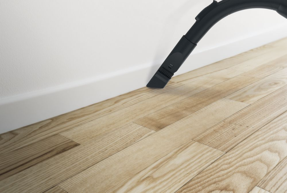 Eine Staubsaugerdüse reinigt einen Holzboden in der Nähe einer weißen Wand und Fußleiste.