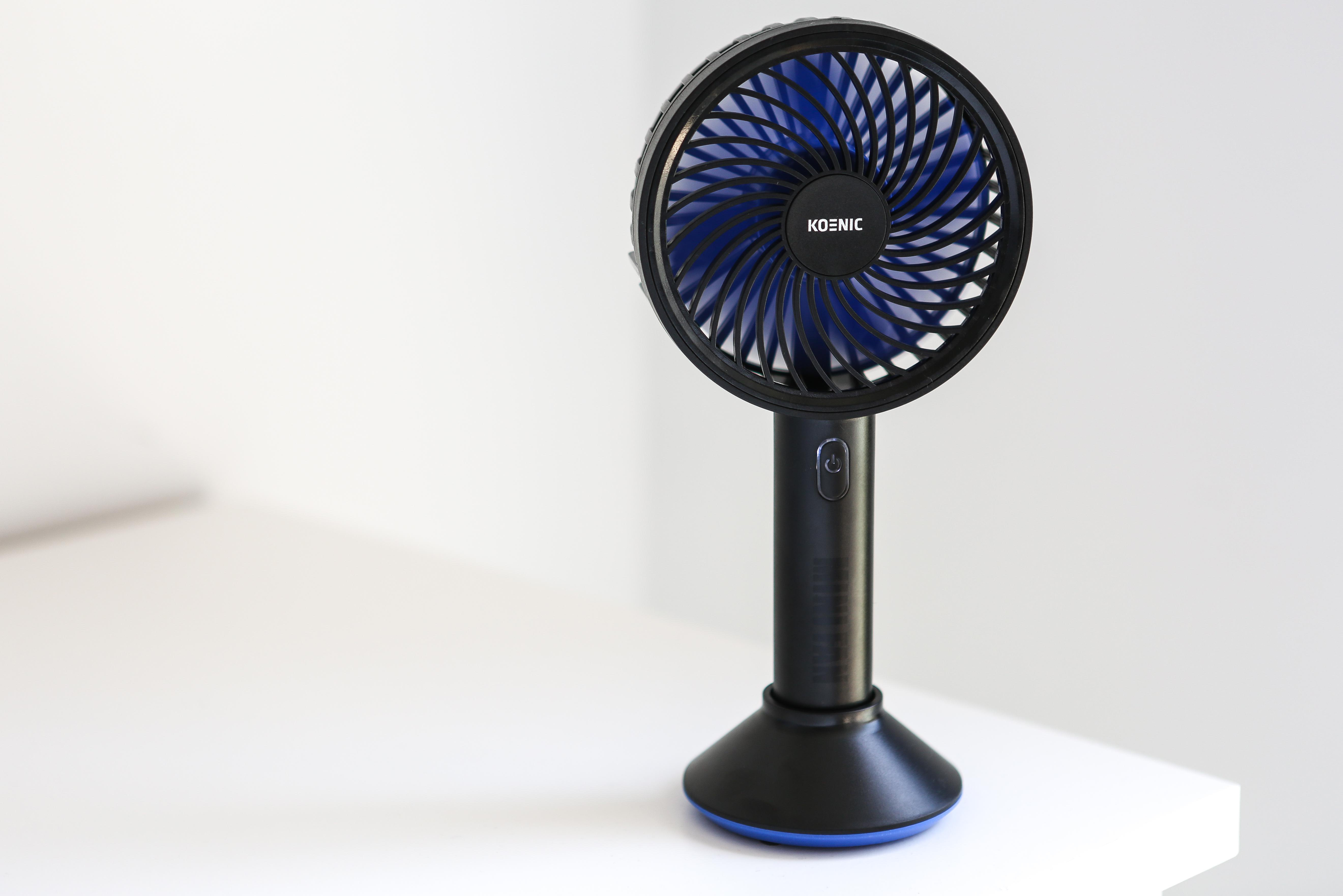 Schwarzer und blauer tragbarer Ventilator auf einer weißen Oberfläche. Er hat einen kreisförmigen Ventilatorkopf und einen schwarzen Sockel.