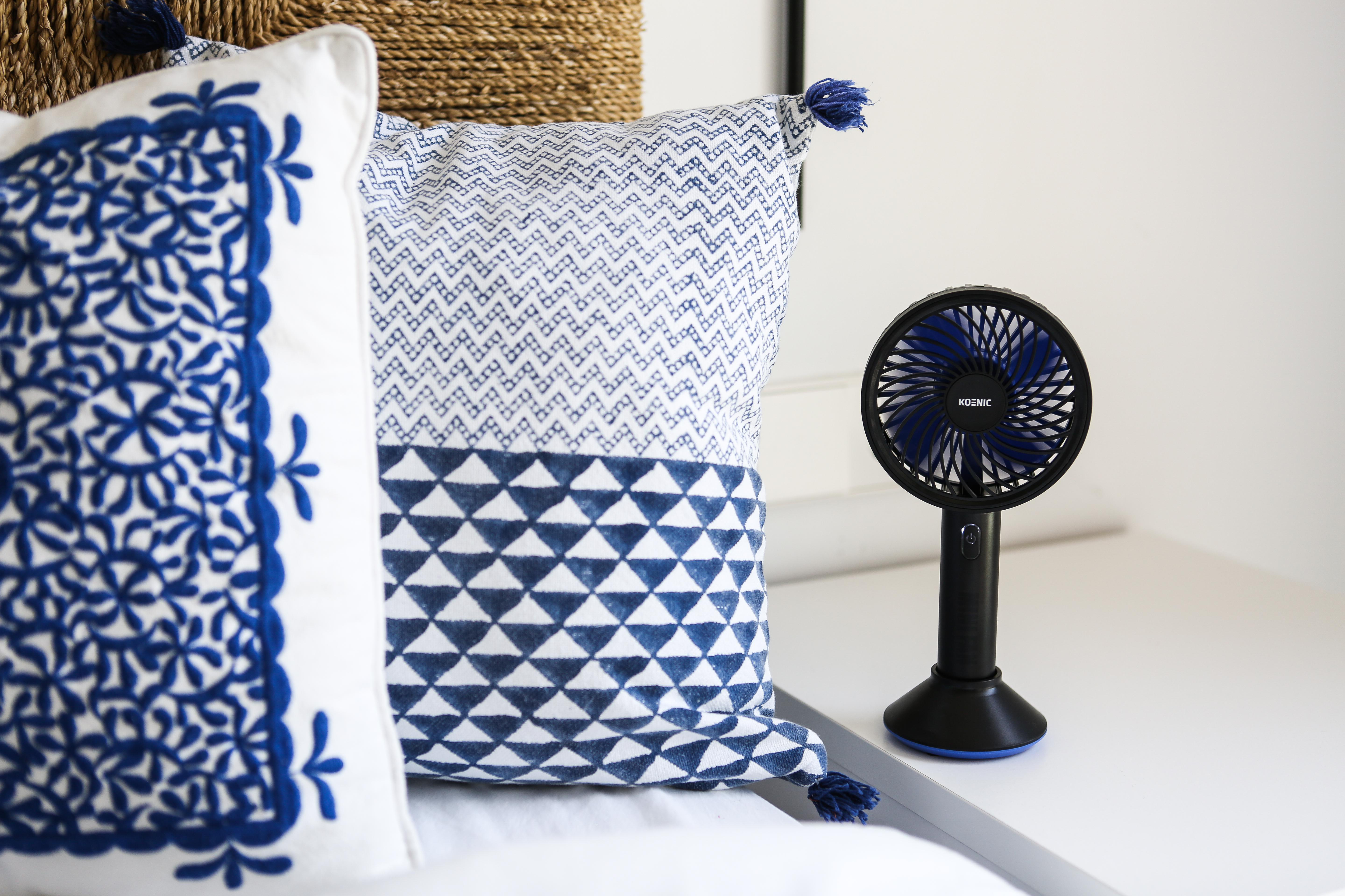 Zwei blau gemusterte Kissen, ein geflochtener Korb und ein kleiner schwarzer Ventilator auf weißer Oberfläche.