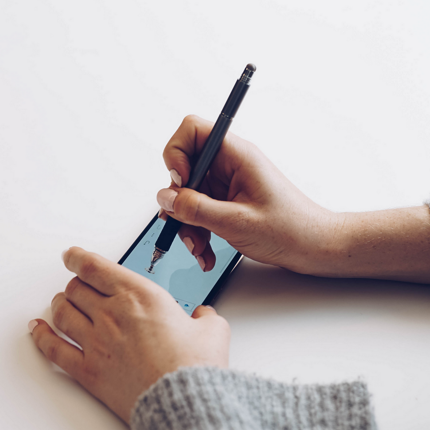 Hands holding a stylus over a smartphone. The screen shows an app. The background is white.