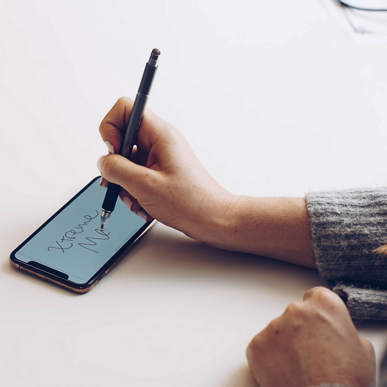 A person's hand holding a stylus on a phone, with text on the screen reading 'Xtreme Me'.