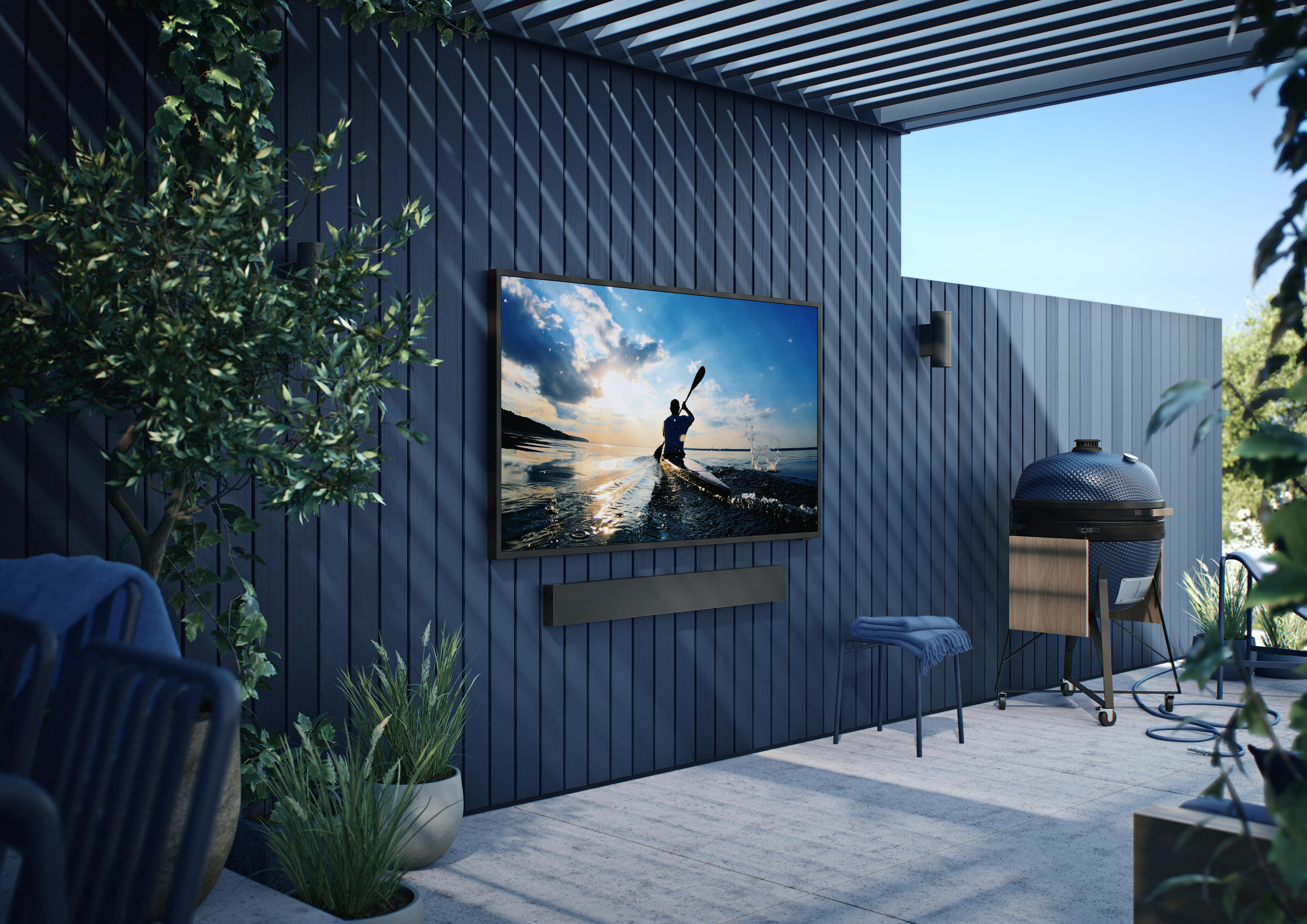 Szene im Freien mit einem Fernseher an einer blauen Holzwand. Ein Grill und Pflanzen sind ebenfalls vorhanden.