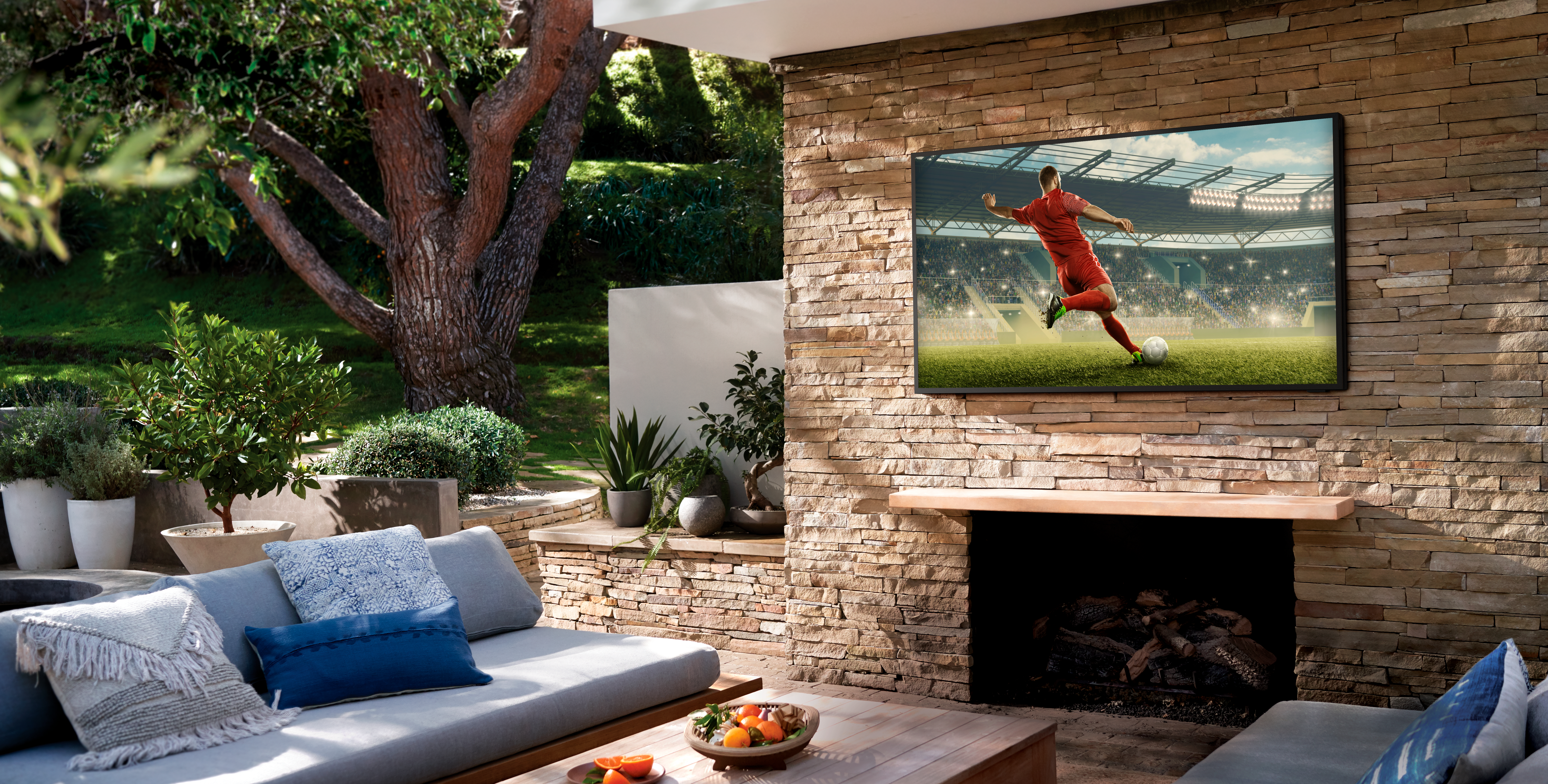 Szene im Freien: Ein Fernseher an einer Steinmauer, der ein Fußballspiel zeigt. Eine Couch und Pflanzen sind zu sehen.
