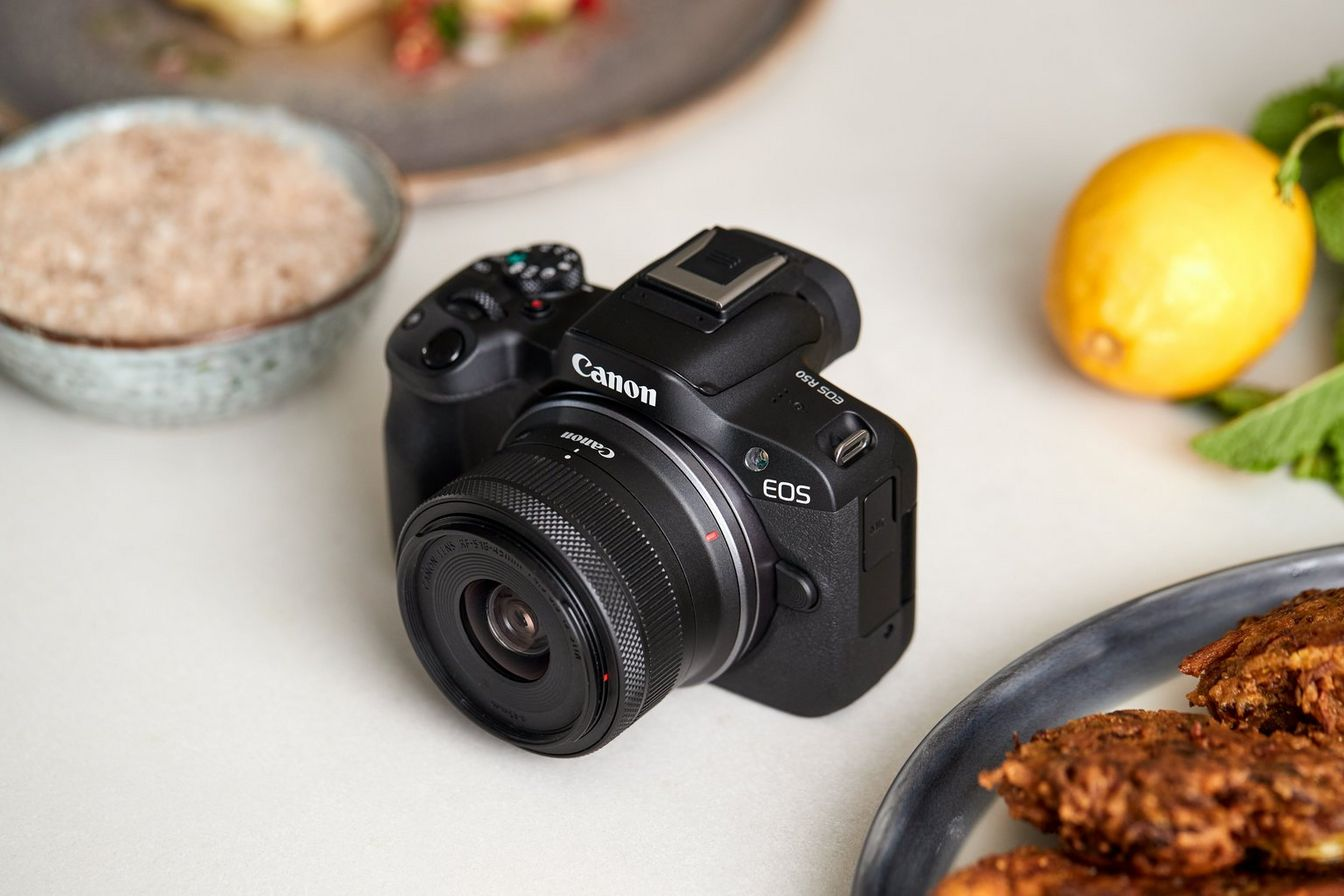 Bir lensle Canon EOS kamera, beyaz bir yüzeyde yiyeceklerle çevrili, iyi aydınlatılmış.