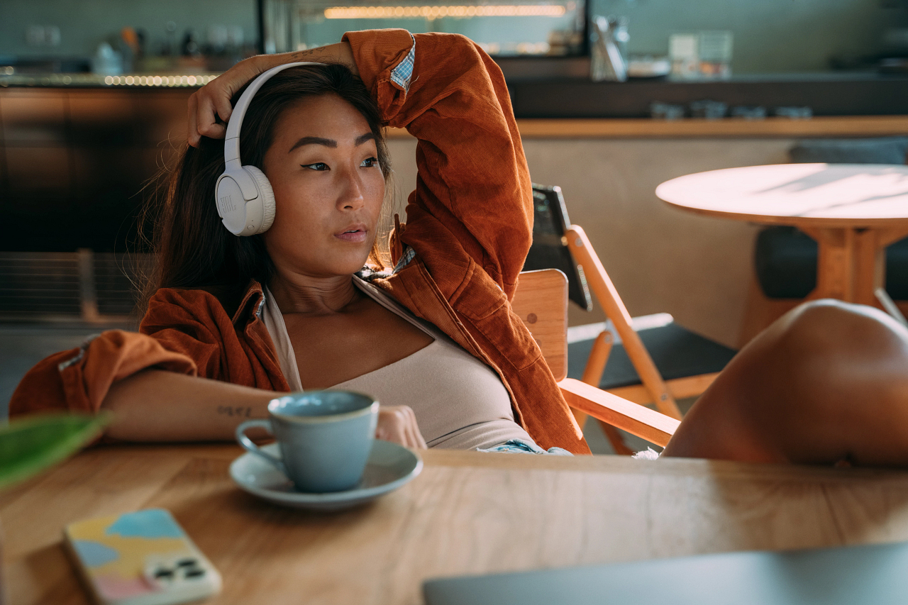 Asiatische Frau mit Kopfhörern, lehnt am Tisch. Kaffeetasse und Telefon daneben.
