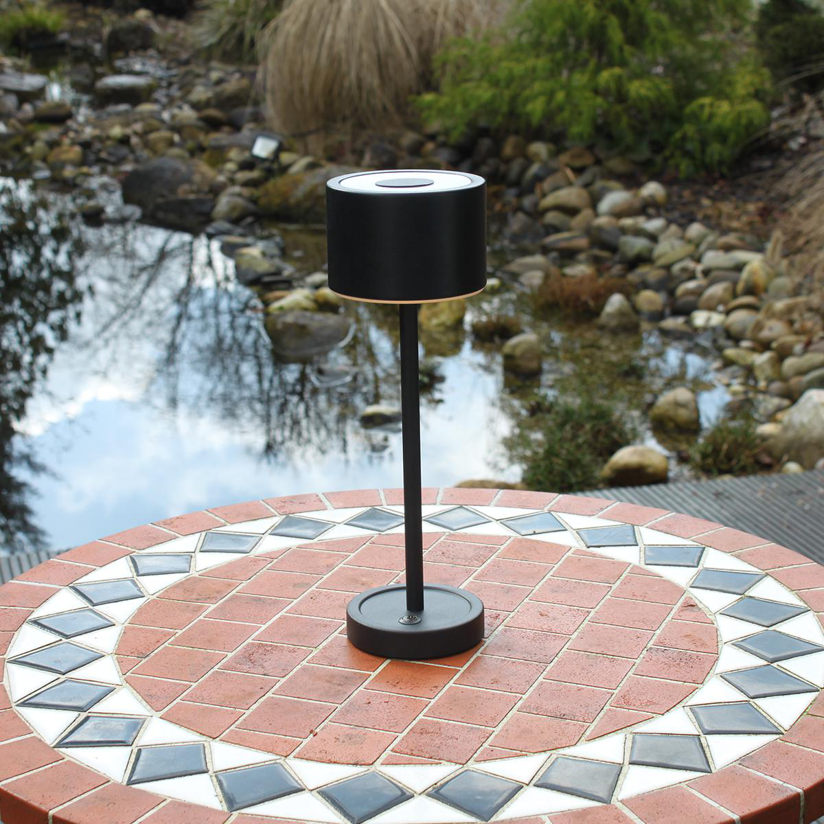 Schwarze Außenlampe auf einem runden Tisch mit Mosaikoberfläche, Wasser und Bäume im Hintergrund.