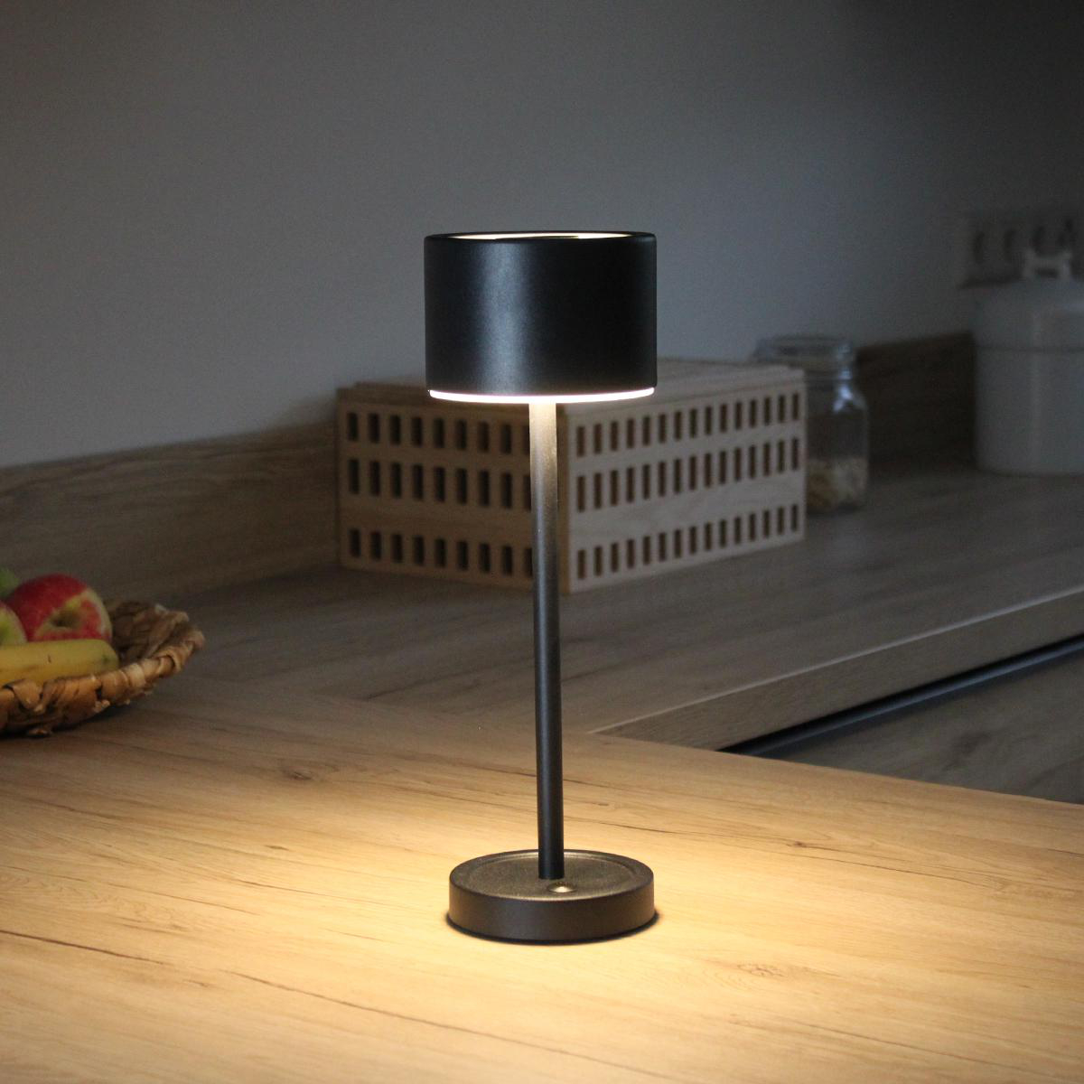 Eine schwarze Tischlampe mit zylindrischem Schirm, auf einer Holz-Arbeitsplatte, warmes Licht.