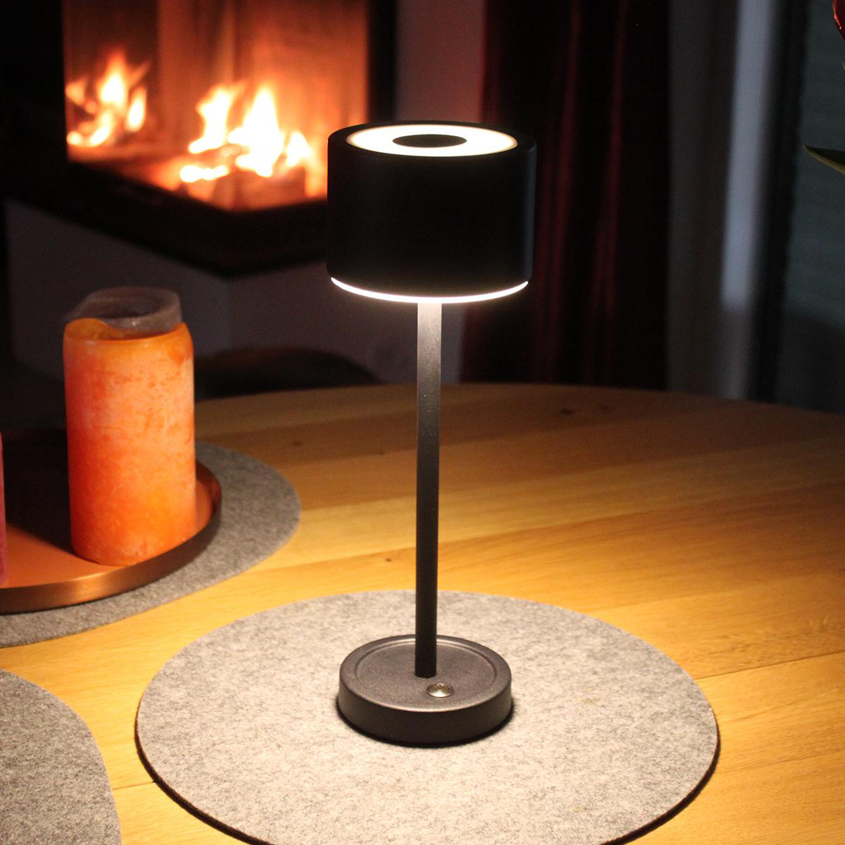 Schwarze zylindrische Tischlampe mit beleuchteter, runder Oberseite, auf einer runden grauen Matte auf einem Holztisch.