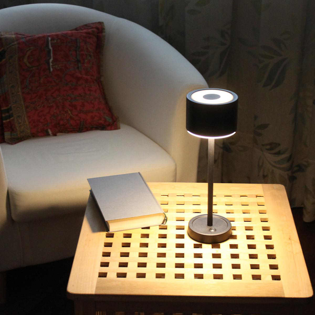 Eine Lampe auf einem Holztisch neben einem Buch und einem weißen Sessel mit rotem Kissen.