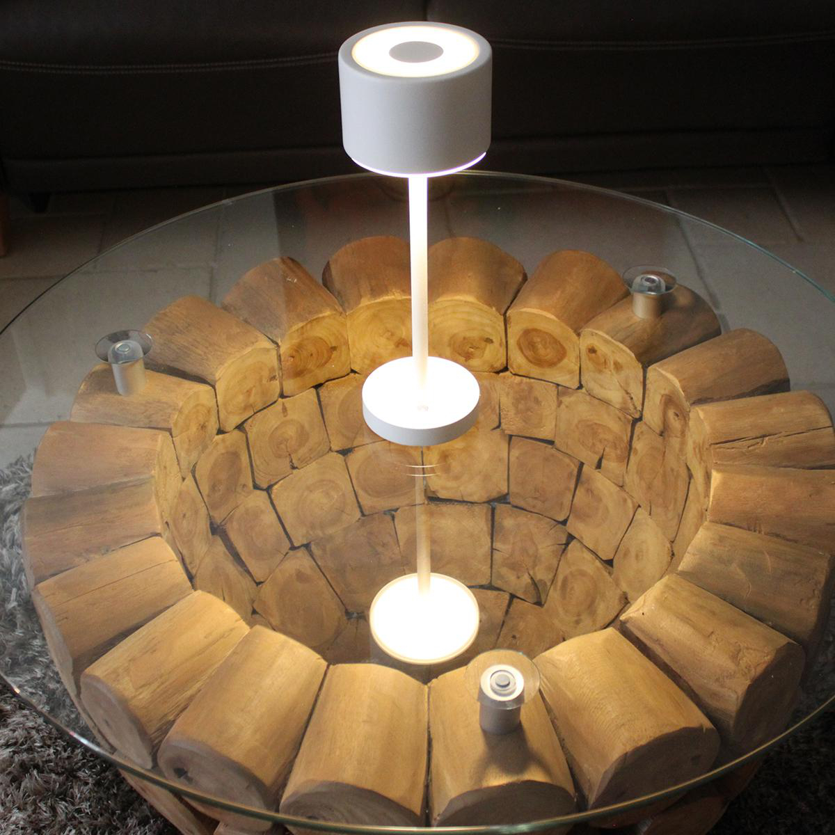 Eine Lampe über einem Holzstamm-Tisch mit Glasplatte. Spiegelung der Lampe sichtbar.