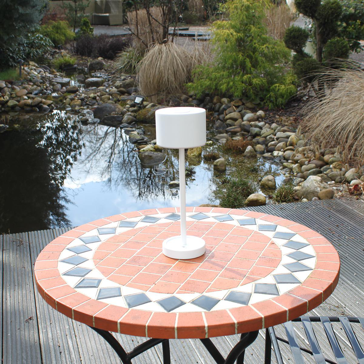 Eine weiße Lampe steht auf einem Tisch, mit einem Teich und Pflanzen im Hintergrund.