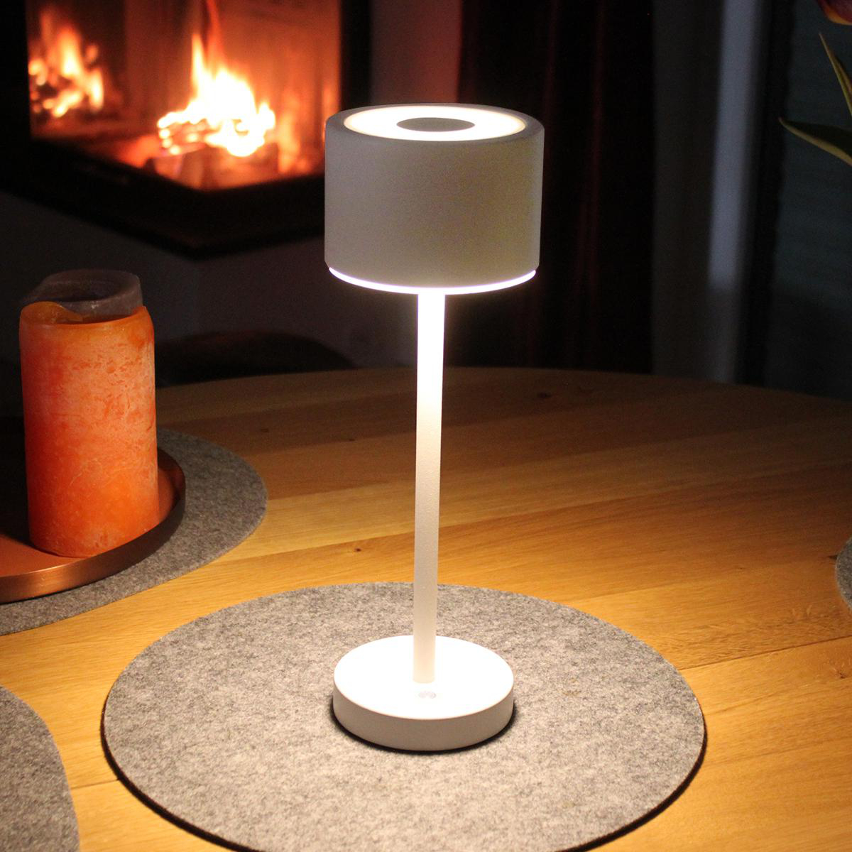 Weiße Tischlampe mit zylindrischem Schirm auf einem Tisch mit grauem Platzset, Kamin im Hintergrund.
