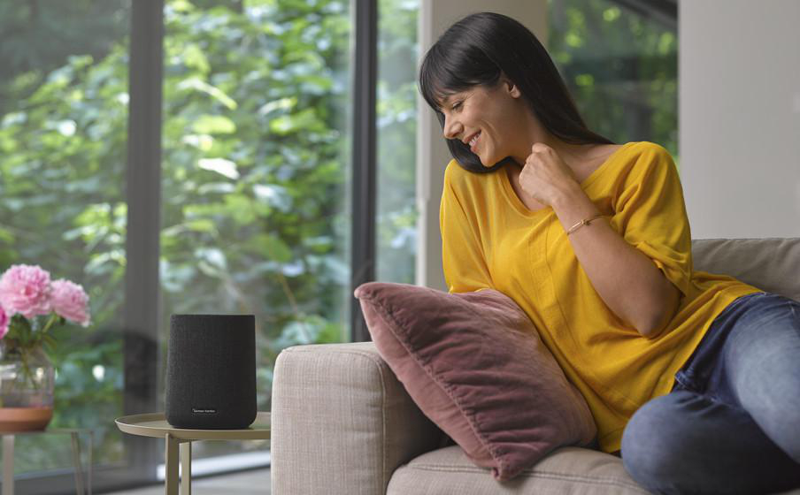 Frau auf Couch mit Smart Speaker und Fenster im Hintergrund.