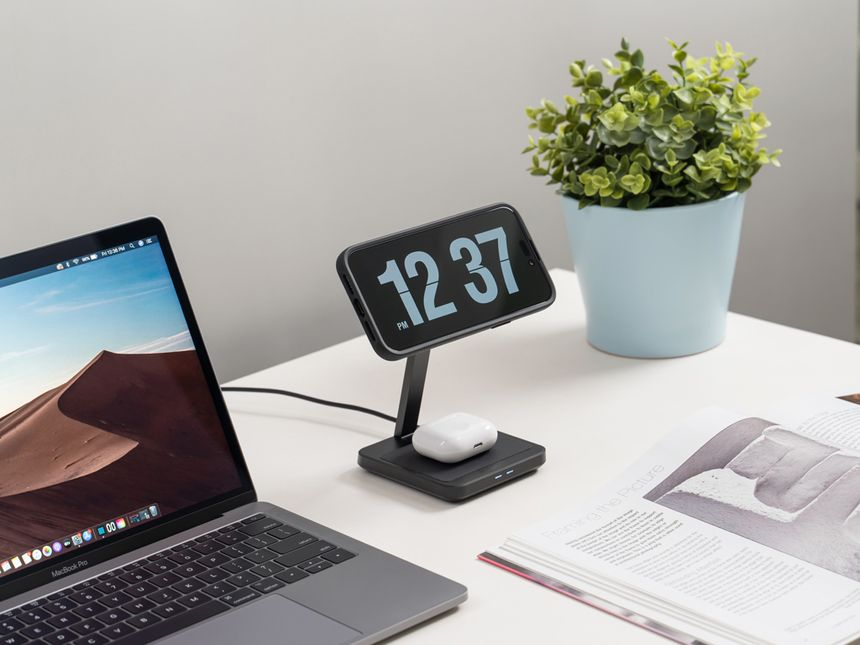 Un'impostazione da scrivania con un laptop, un orologio, auricolari wireless e una pianta in un vaso blu.