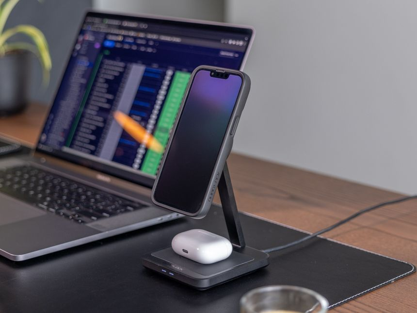 Un'impostazione da scrivania con un laptop, un telefono su un caricabatterie e una custodia per airpod bianca.