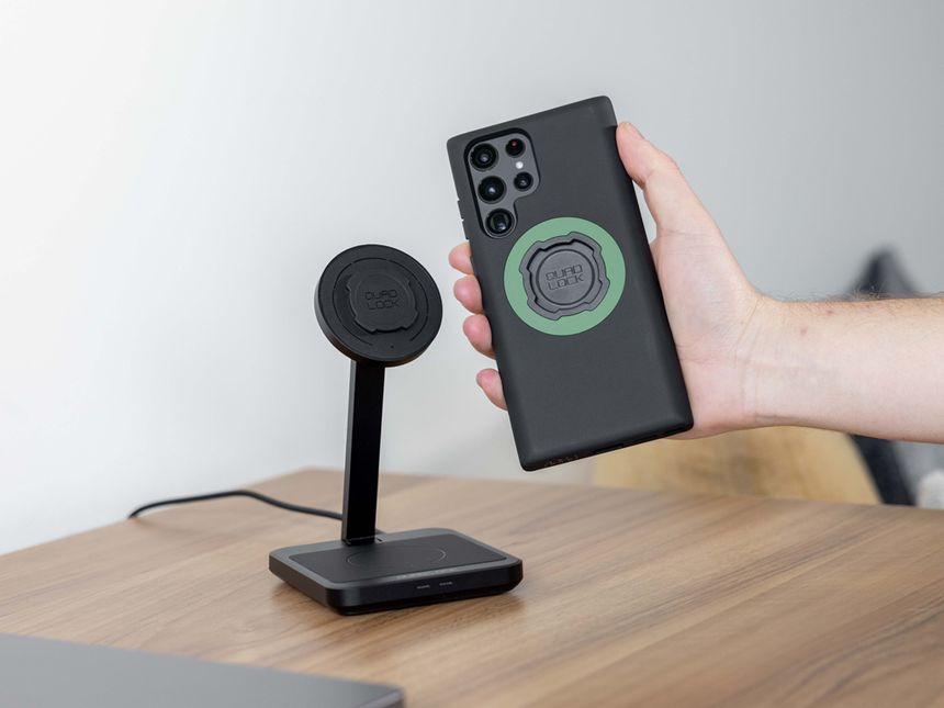 Mano che tiene un telefono nero con un anello verde su un caricatore wireless. Supporto nero su tavolo di legno.