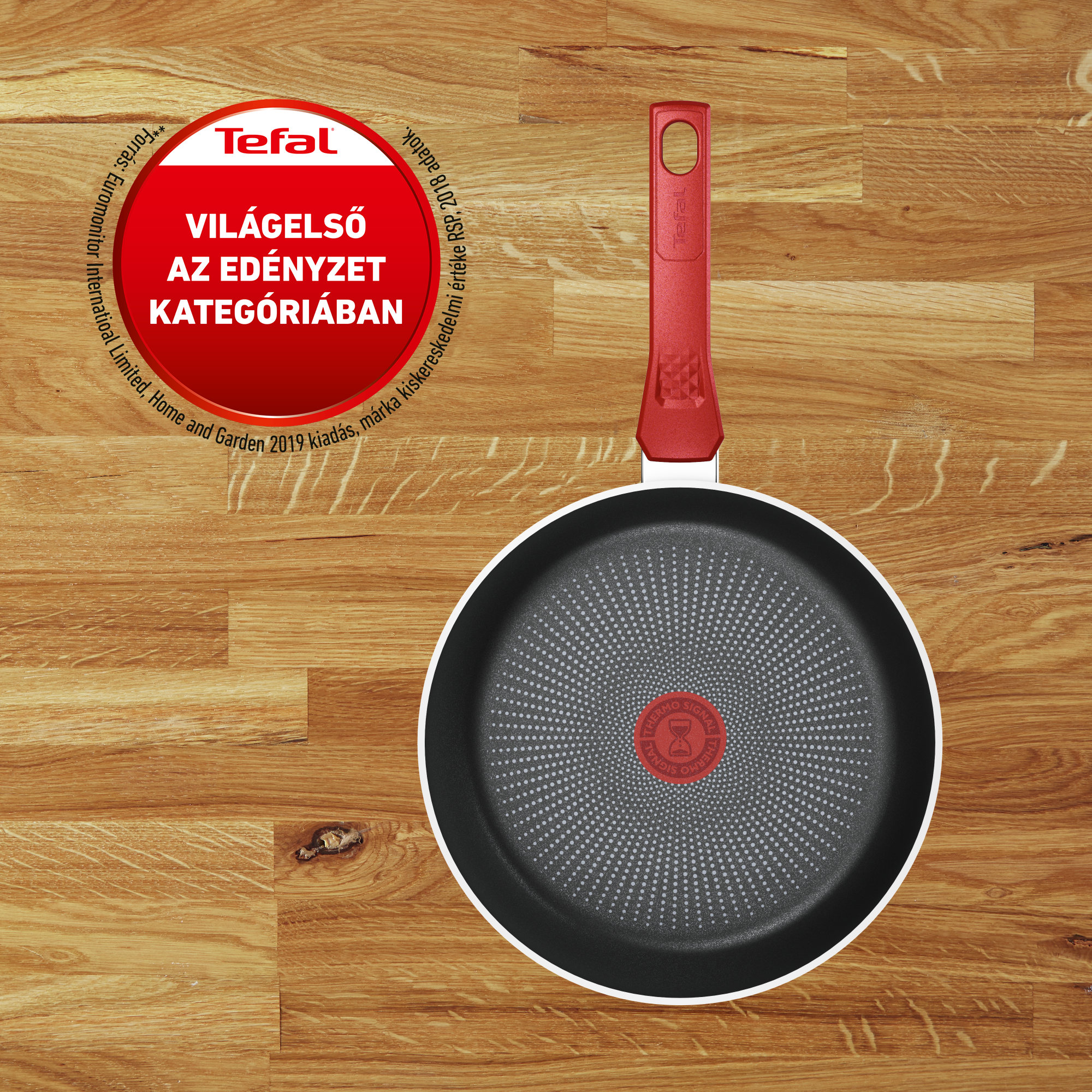 Tefal serpenyő piros fogantyúval fa felületen. A serpenyő fekete, ezüst szegéllyel.