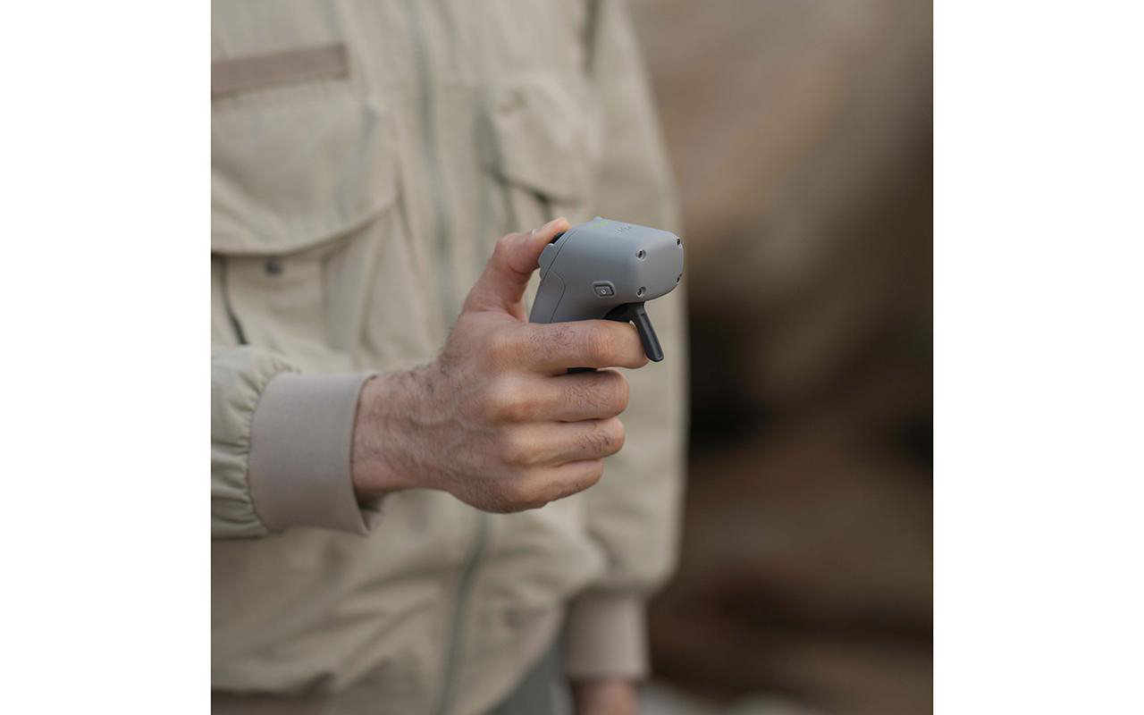 Eine Hand hält eine graue Drohnen-Fernbedienung. Die Person trägt eine beige Jacke, verschwommener Hintergrund.