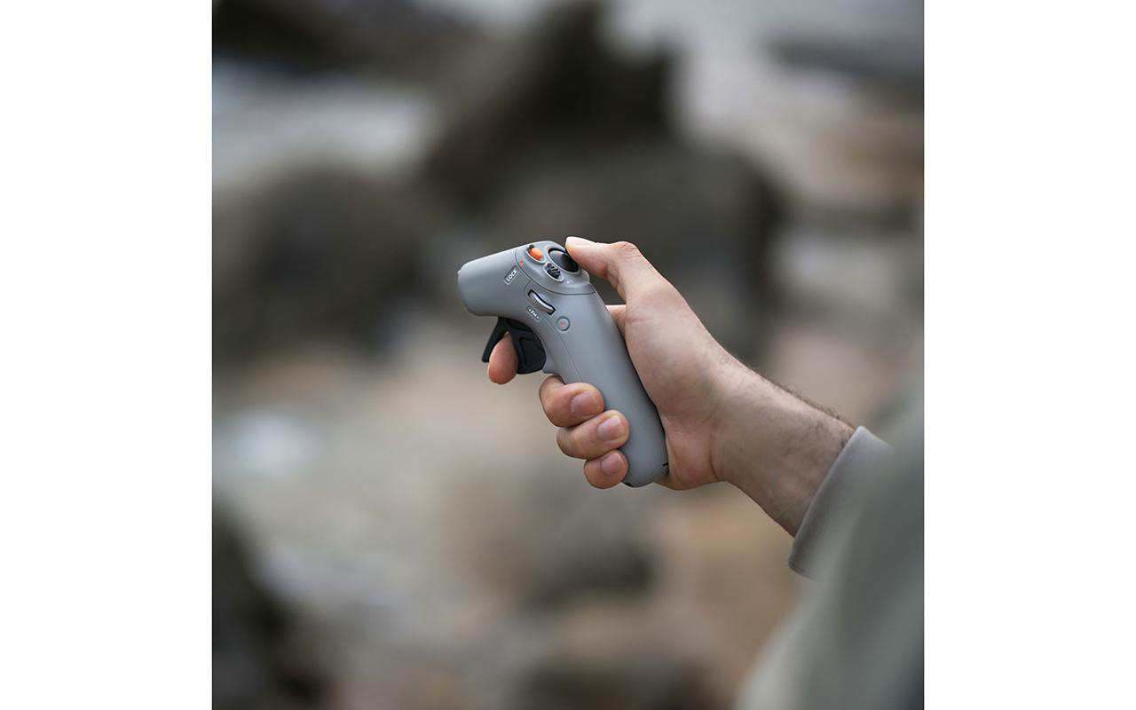 Eine Hand, die einen grauen Controller mit Knöpfen und einem Auslöser hält. Der Hintergrund sind verschwommene Felsen.