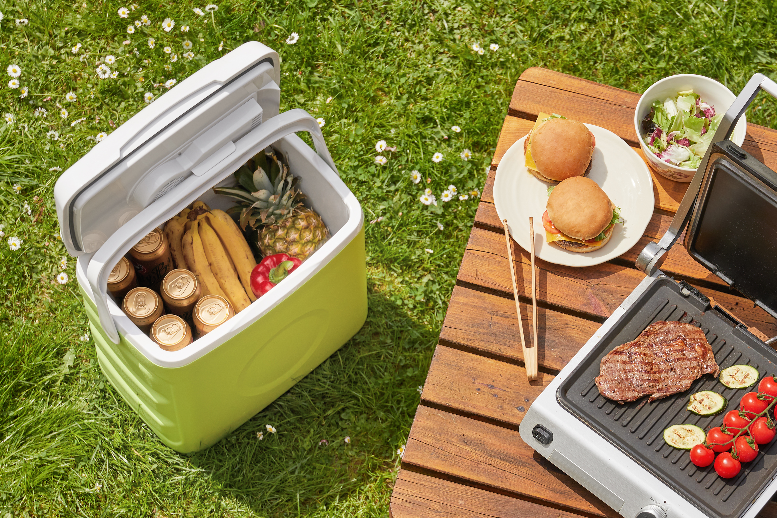 Piknik jelenet: Hűtőtáska, étel és grill a füvön, napos nap.