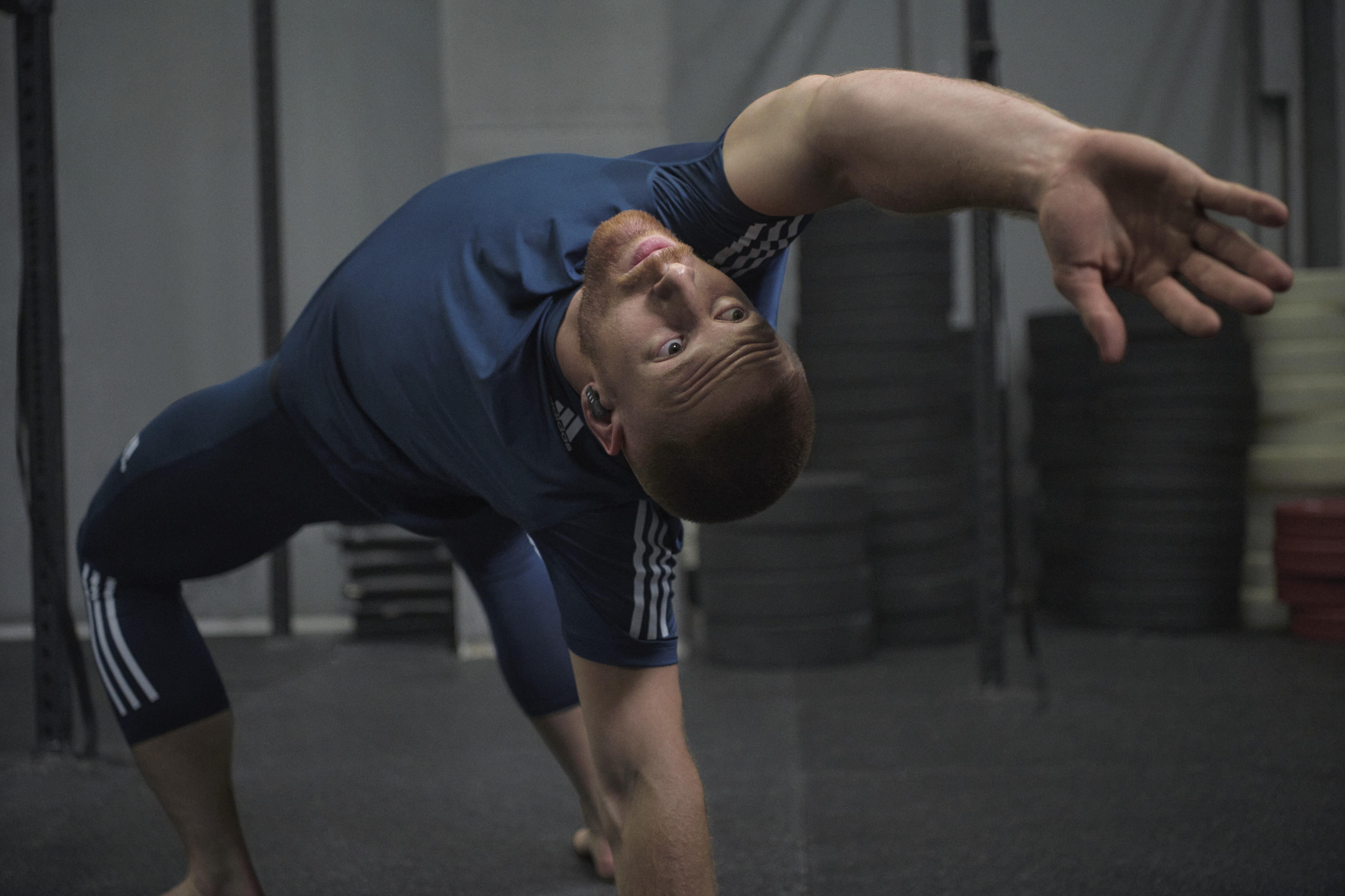 Ein Mann in Sportkleidung beugt sich mit ausgestrecktem Arm in einem Fitnessstudio. Er blickt nach oben.