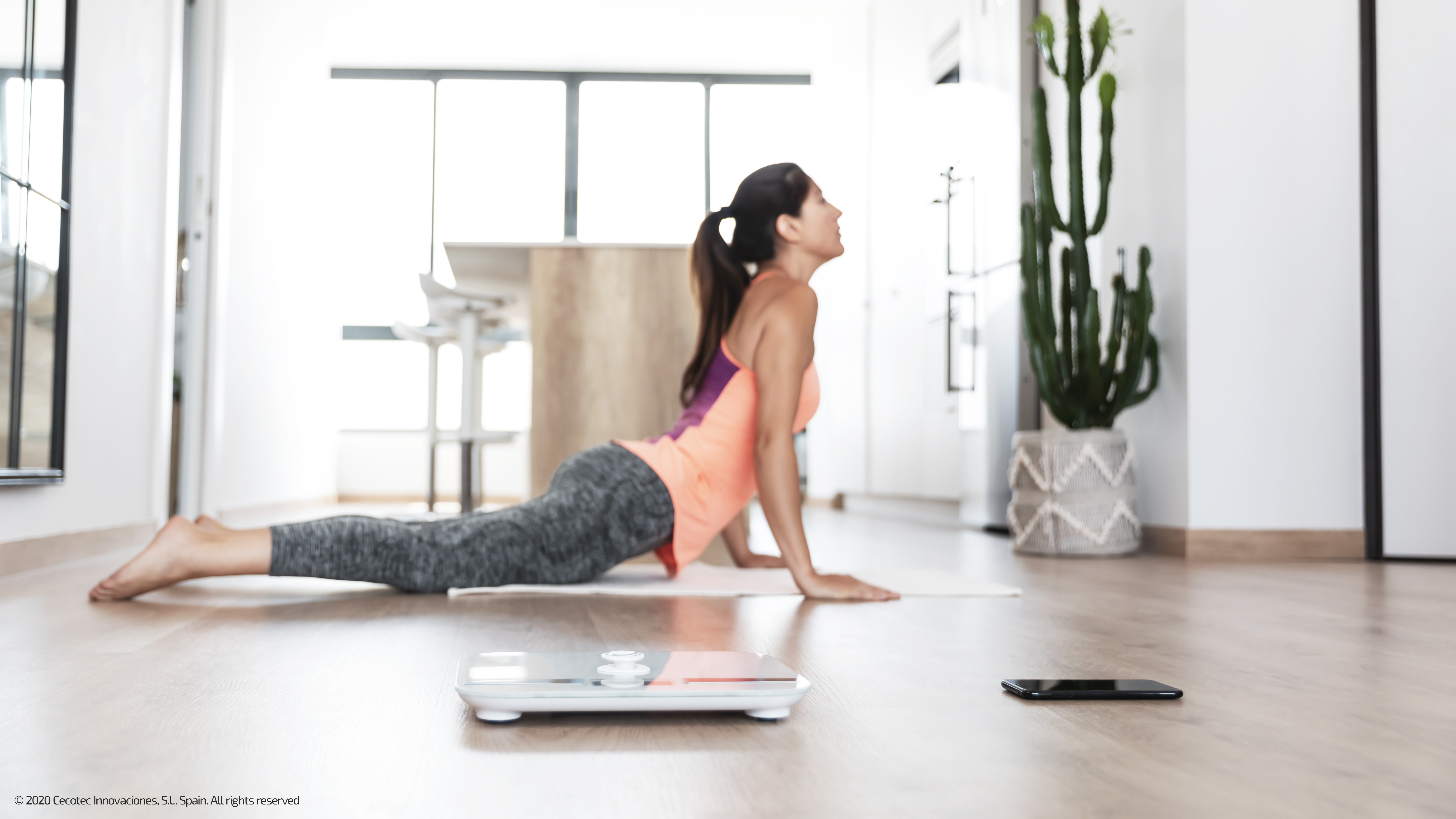 Mujer en postura de yoga en una esterilla, báscula y teléfono cerca en una habitación luminosa.