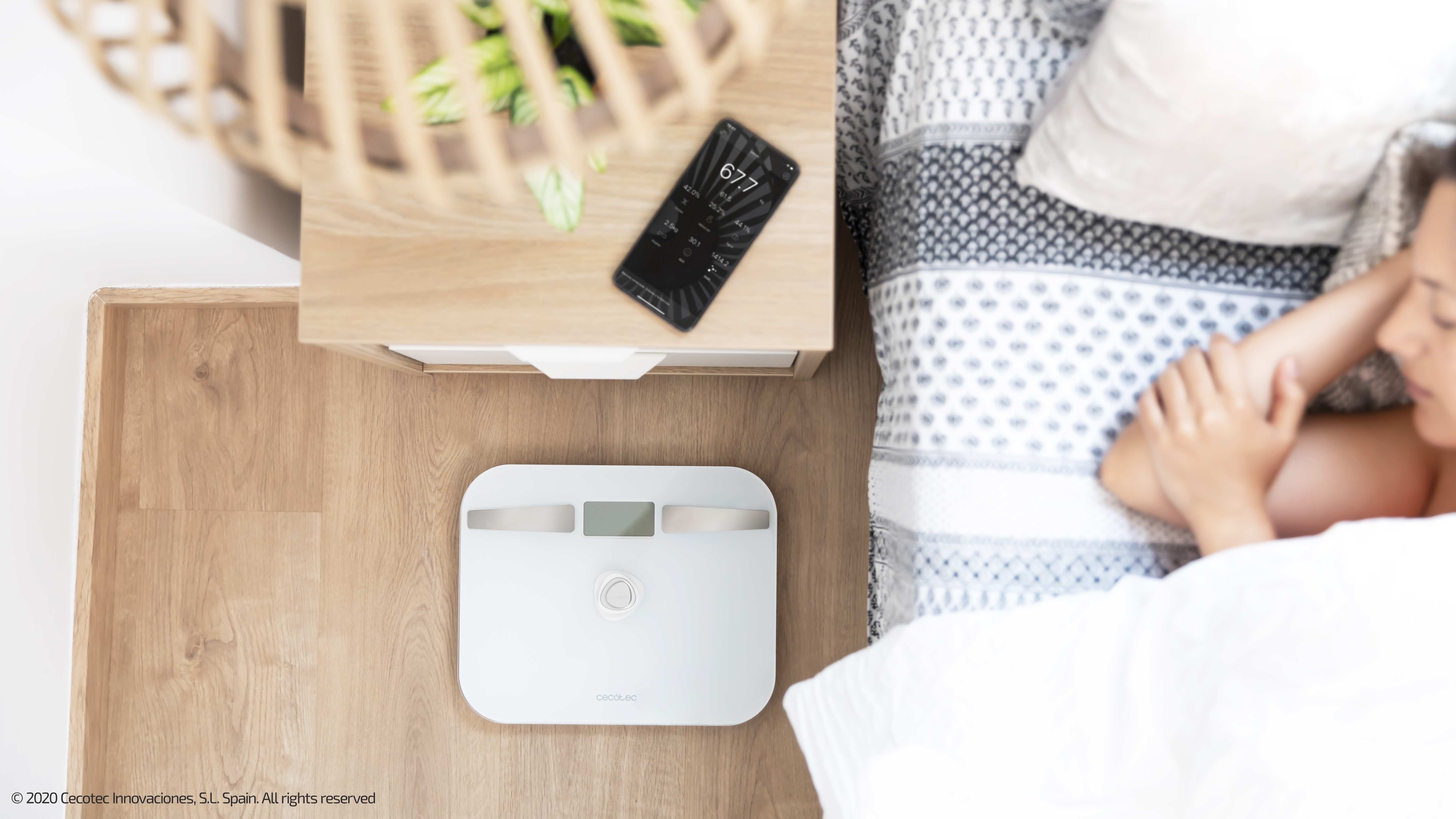 Una mujer descansando, una báscula inteligente en el suelo y un teléfono mostrando datos. Mesita de noche.