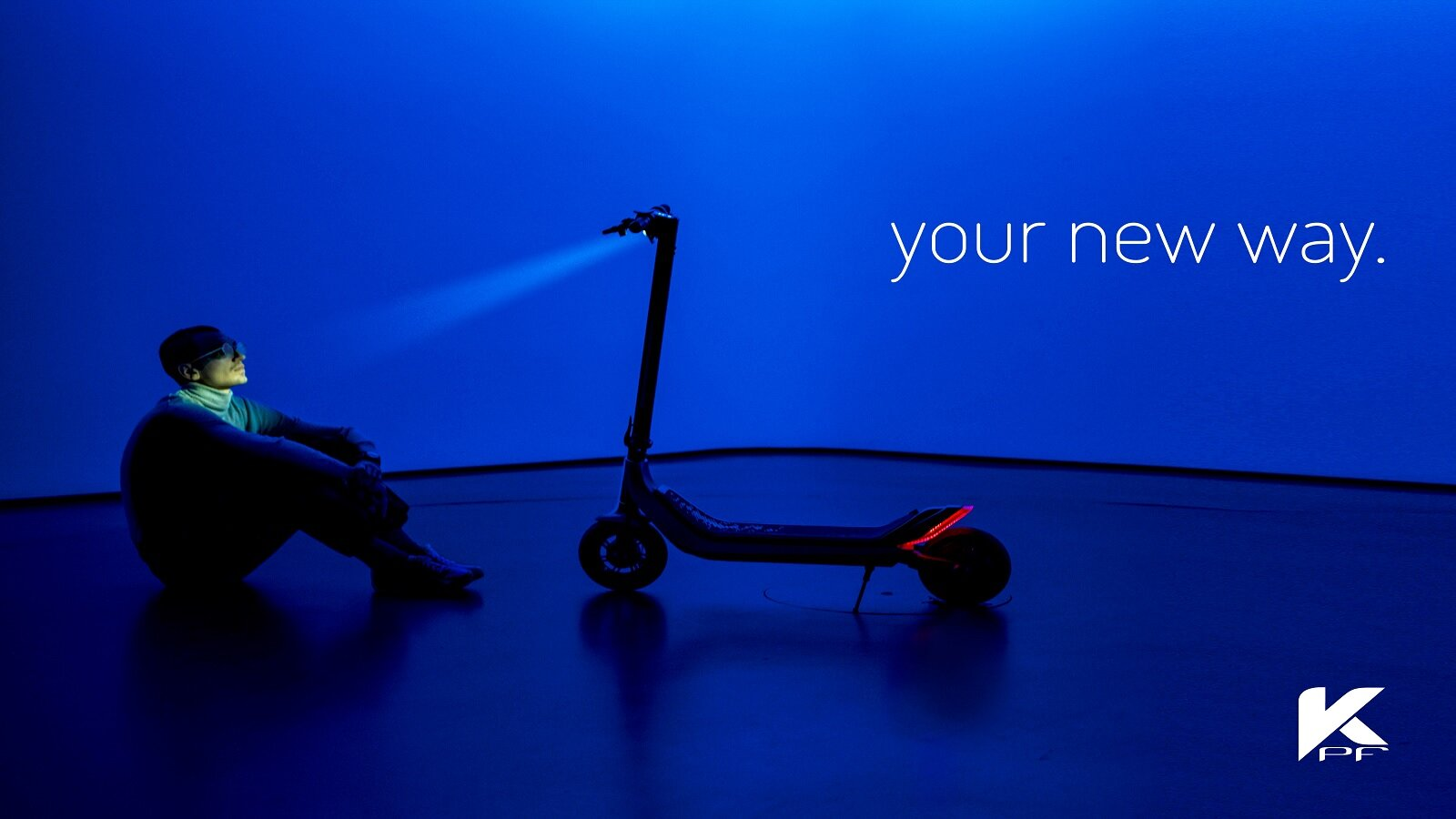 Mężczyzna siedzi obok skutera z reflektorem na niebieskim tle.