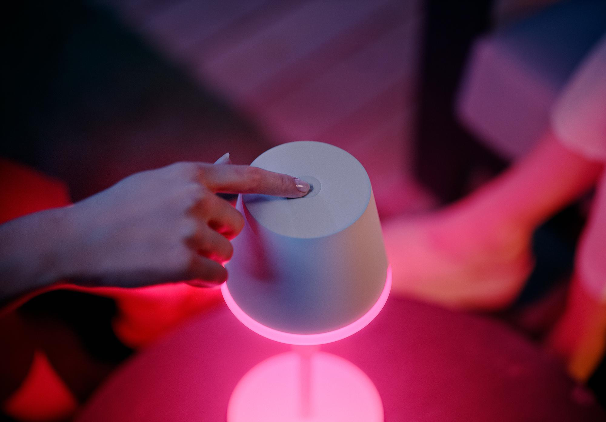 Hand drückt einen Knopf auf einer weißen Lampe, die rosa Licht auf einem Tisch ausstrahlt.