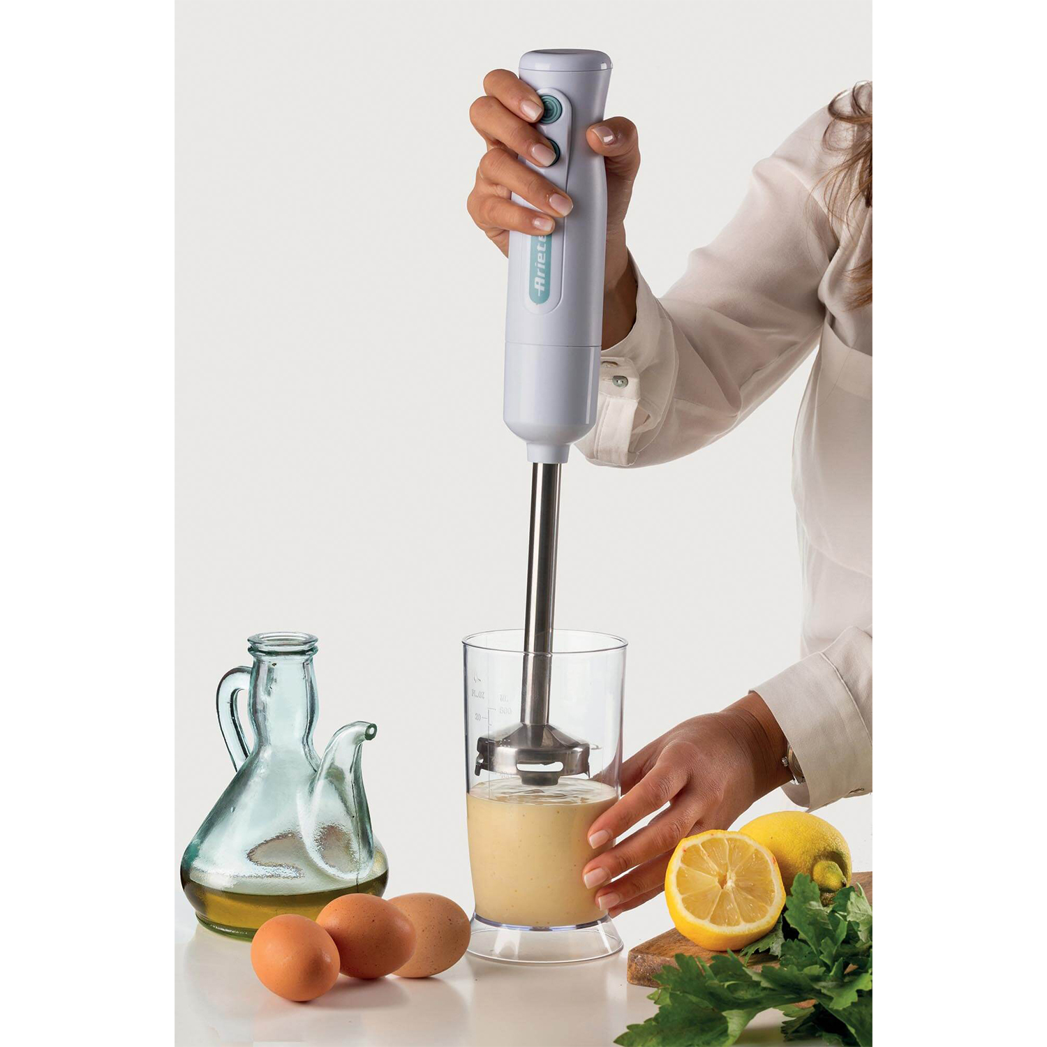 Una persona usa un frullatore a immersione in un bicchiere, mescolando ingredienti. Uova e limone sul tavolo.