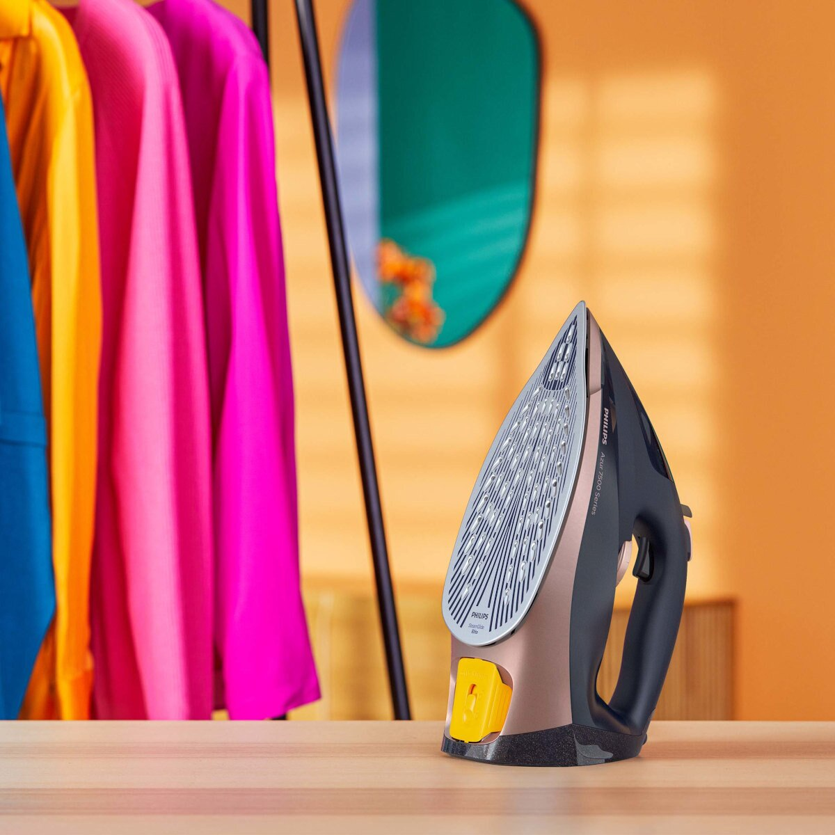 Żelazko na drewnianym stole, z ubraniami i lustrem w tle.