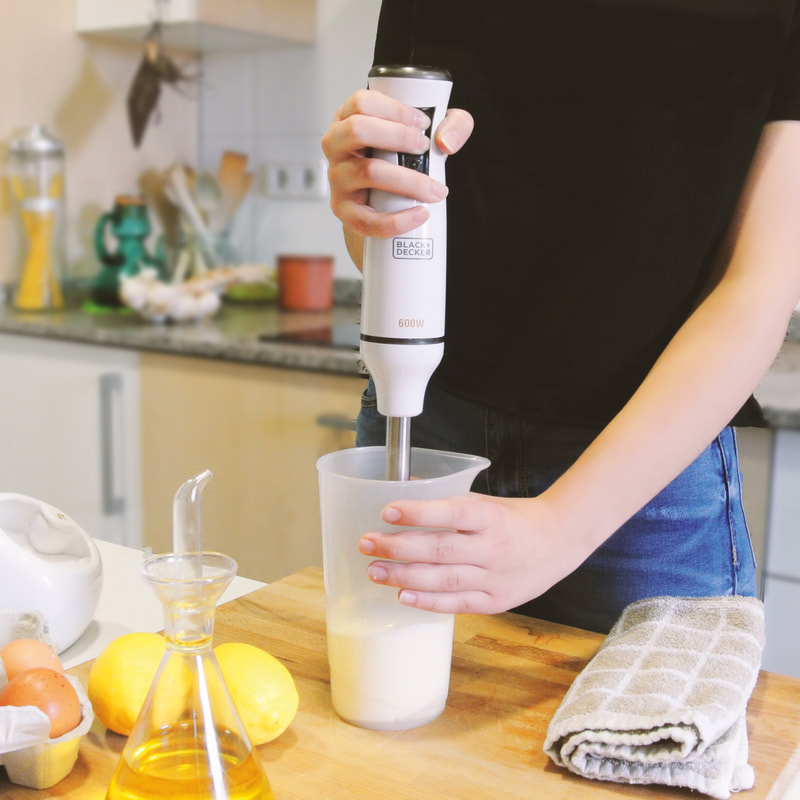 Egy személy kézi mixerrel keveri az összetevőket egy pohárban, olajjal, tojással és citrommal a közelben.