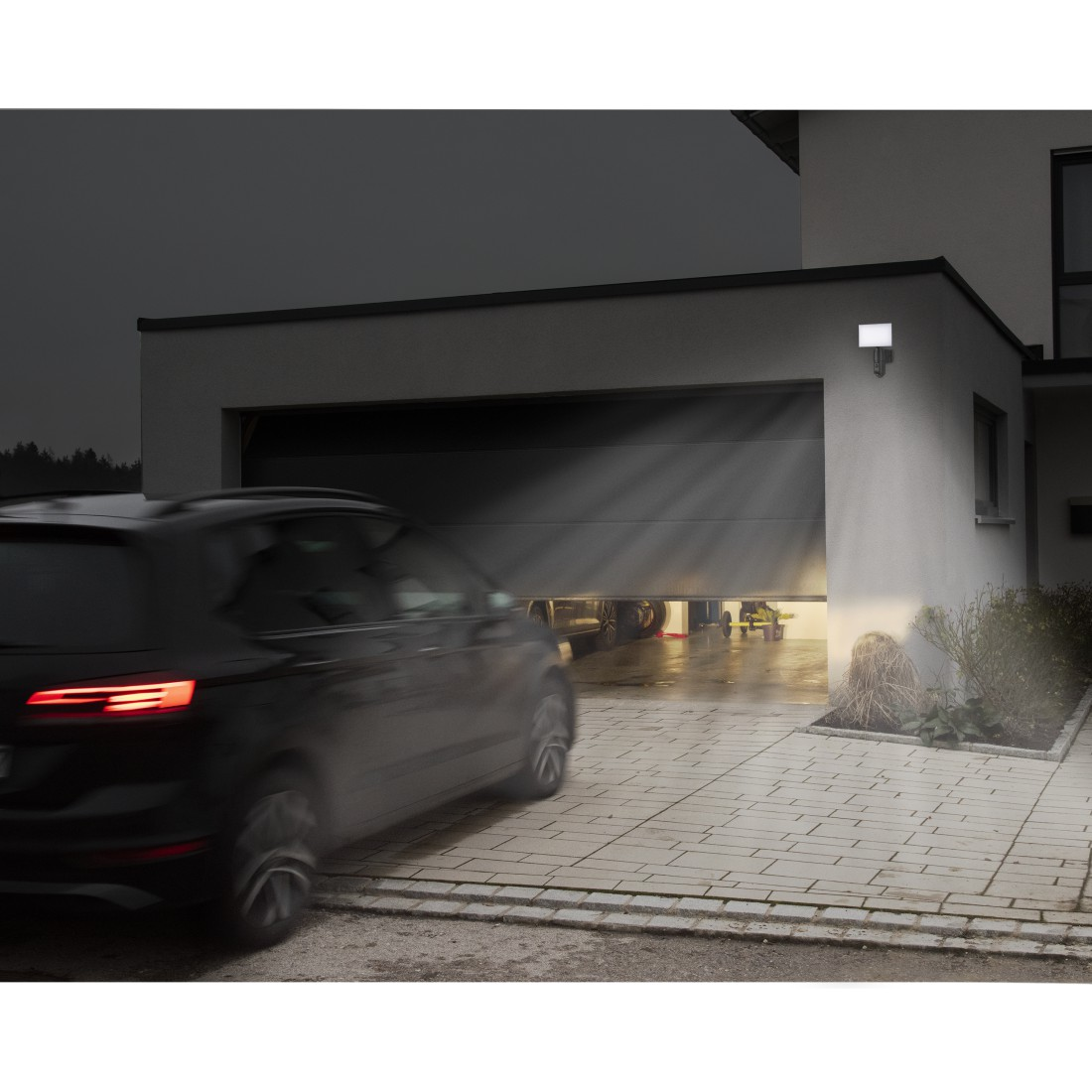 Ein Auto fährt nachts aus einer Garage. Das Garagentor ist offen, und Licht scheint heraus. Ein Scheinwerfer ist an der Wand.