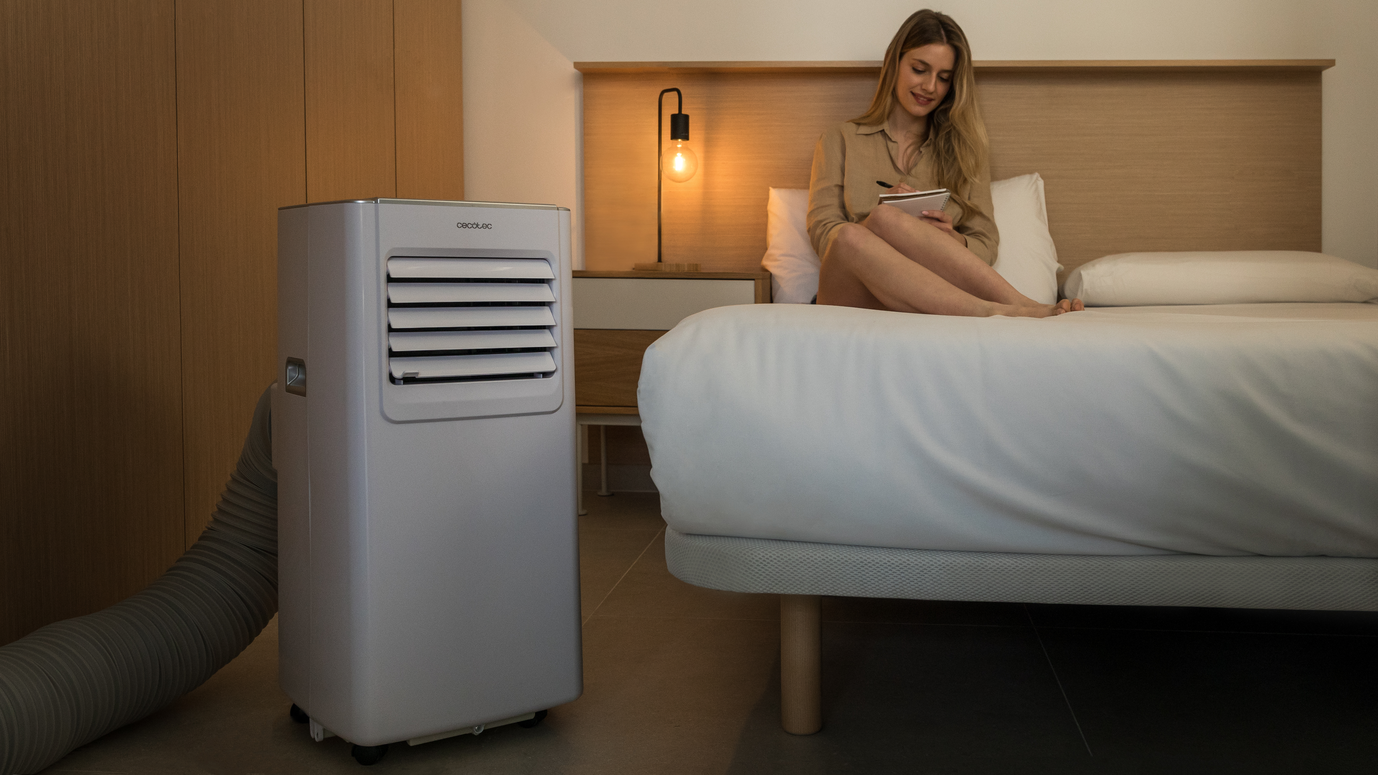 Una mujer escribe sentada en una cama. Una unidad de aire acondicionado blanca junto a la cama. Hay una lámpara y muebles de madera.