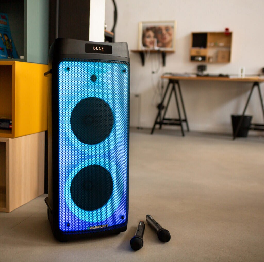 Wysoki czarny głośnik z niebieskim podświetleniem. Dwa mikrofony z przodu. Tło salonu z dziełami sztuki.