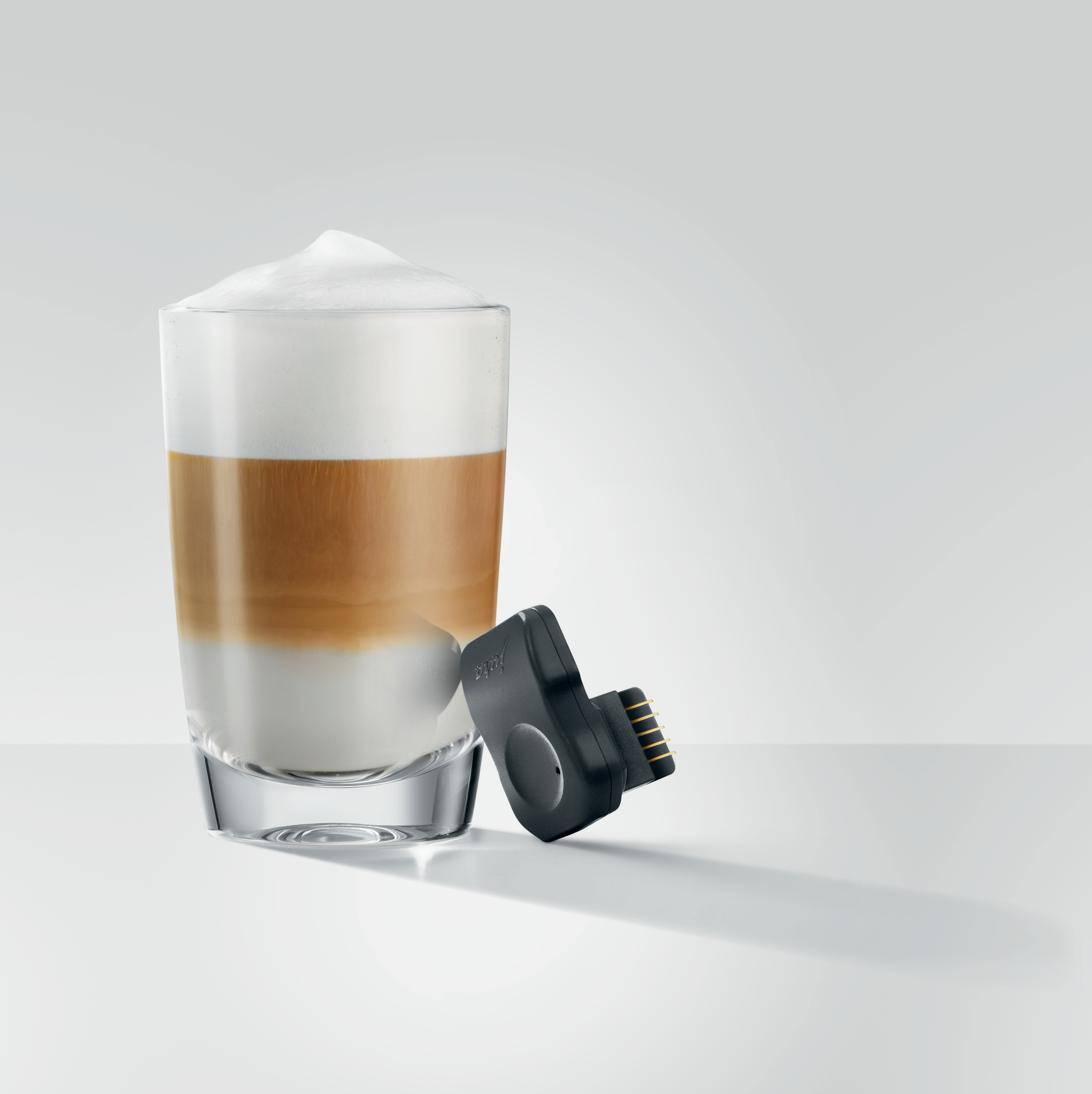 Ein Latte mit Schaum in einem Glas, daneben ein kleines schwarzes Gerät.