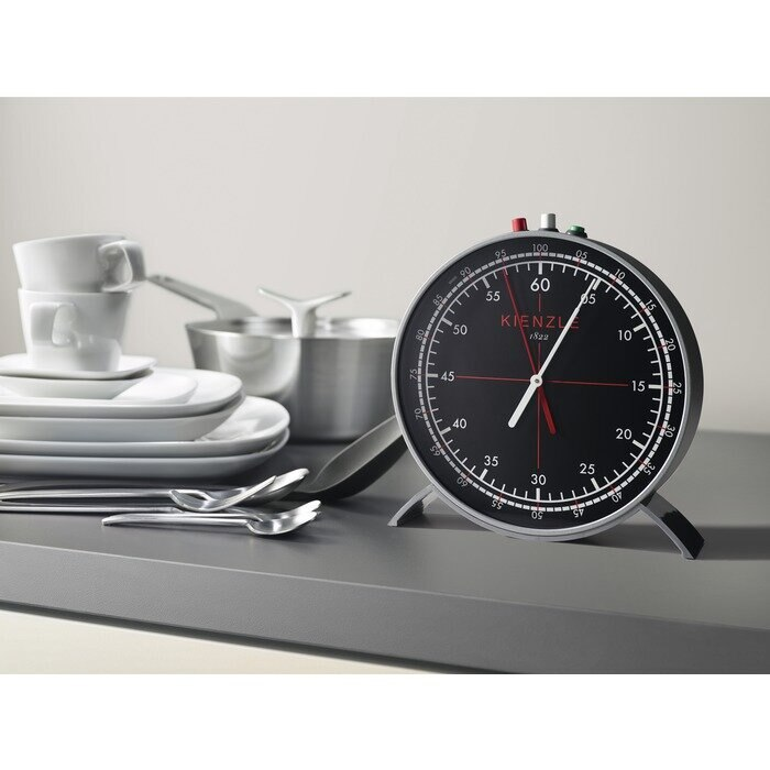 Czarny i srebrny timer Kienzle na szarej powierzchni. Białe naczynia i sztućce w tle.
