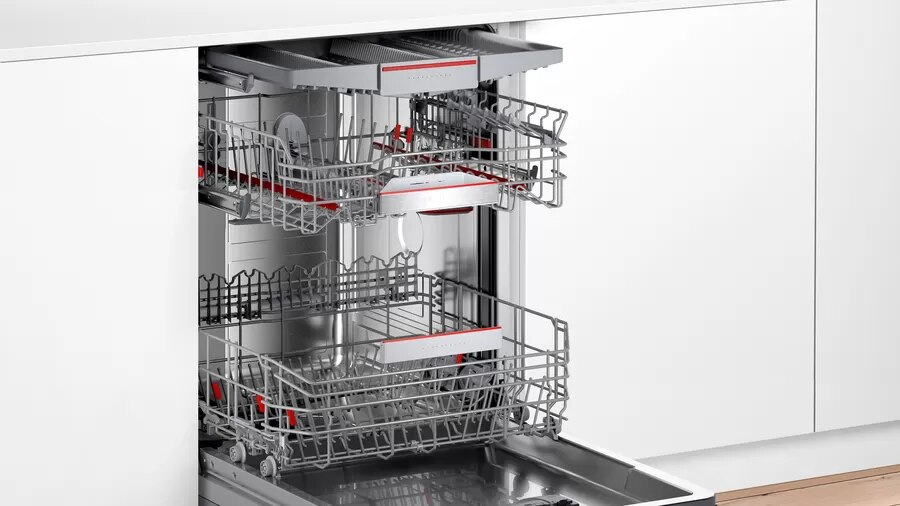 Otwarta zmywarka z dwoma koszami. Kosze puste z czerwonymi akcentami. Białe wnętrze i szafki.