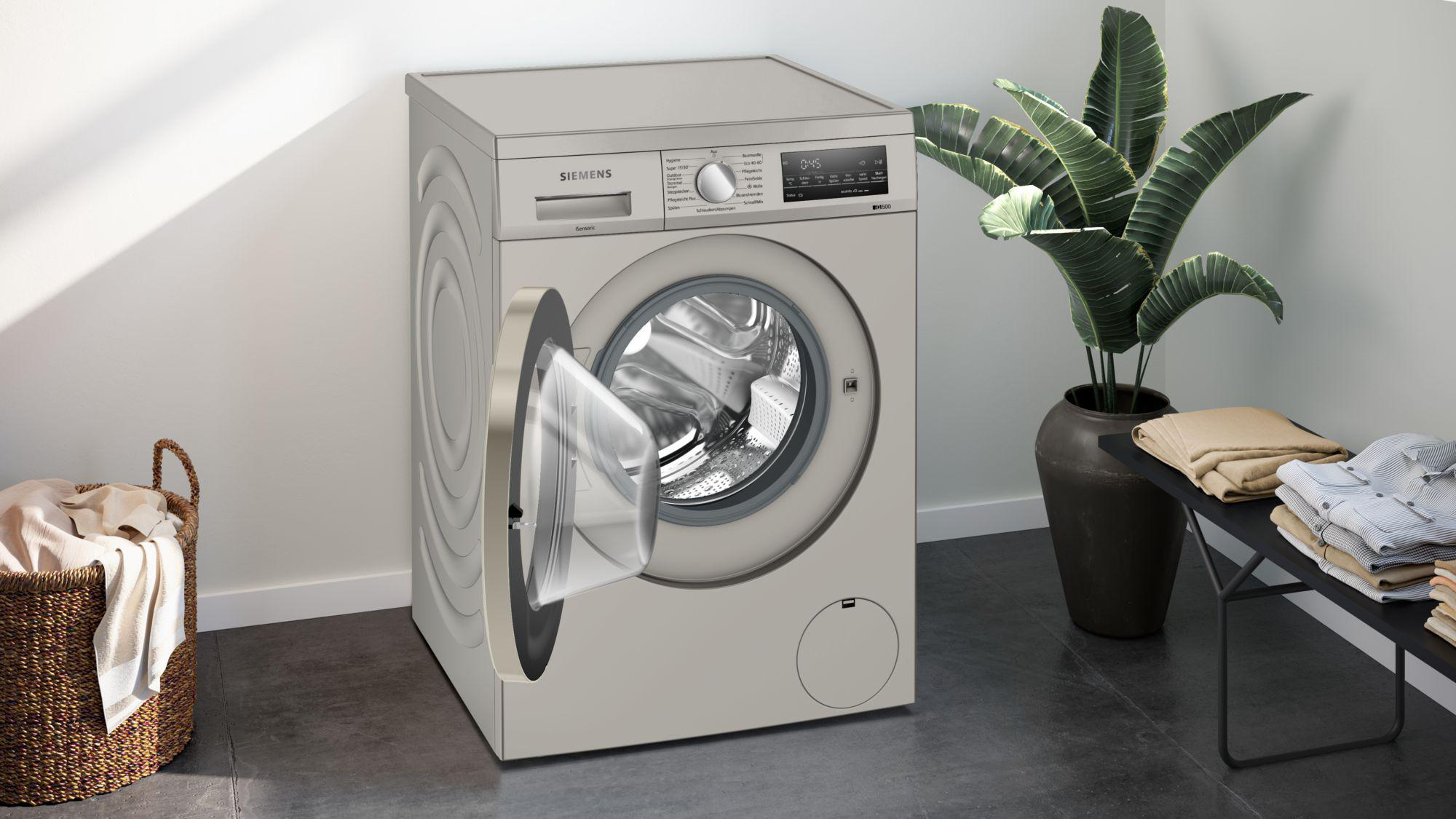 Eine Siemens-Waschmaschine mit offener Tür steht in einem Raum. Rechts befinden sich ein Korb mit Kleidung und eine Pflanze in einem Topf.