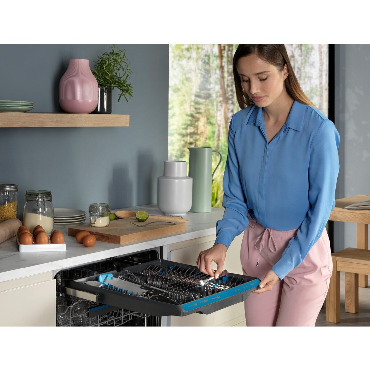 Kobieta ładująca szufladę zmywarki sztućcami, z przedmiotami kuchennymi w tle.