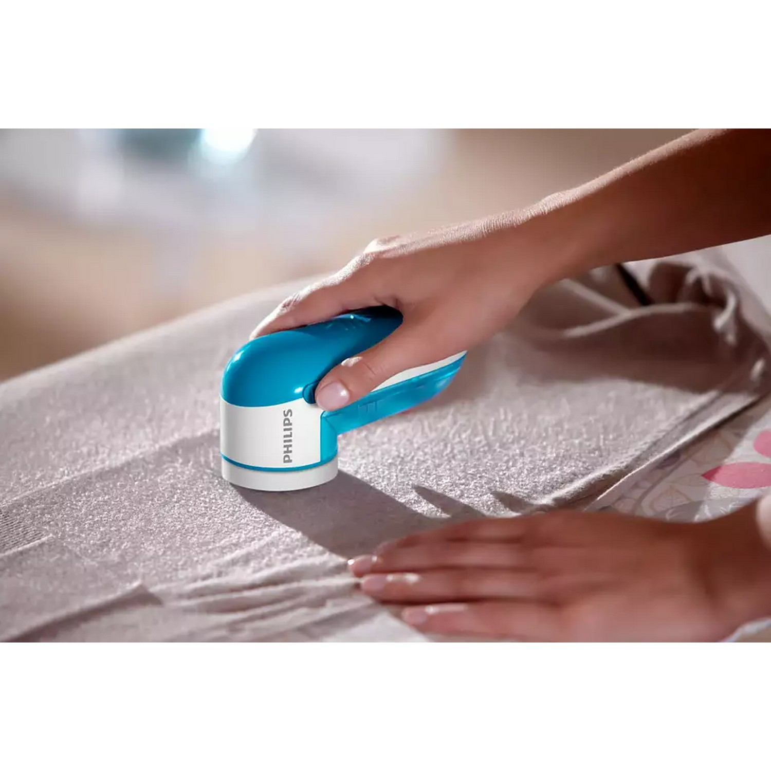 A person uses a blue and white Philips fabric shaver on a gray sweater.
