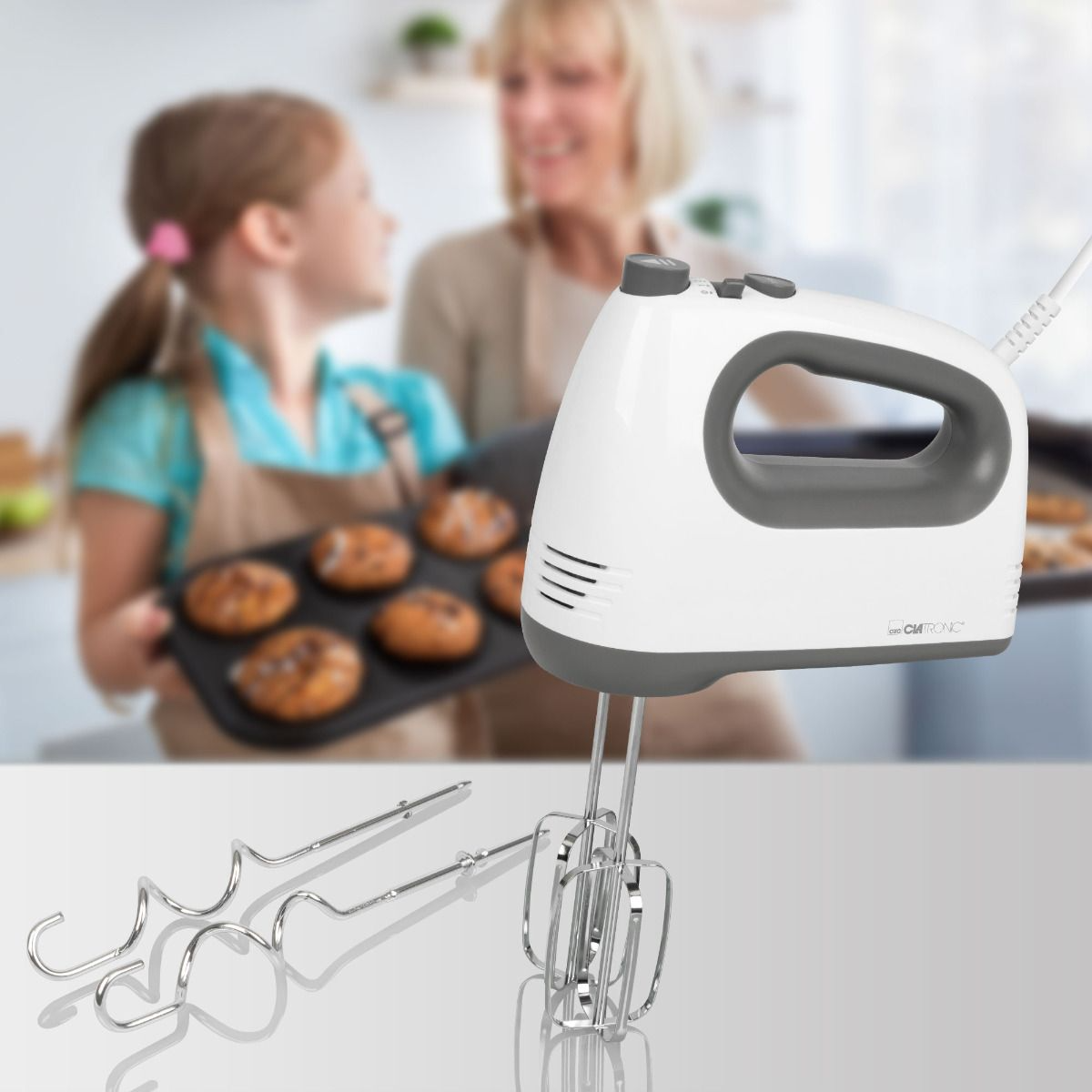 Kézi mixer használatban. Egy kislány tepsit tart, egy nő nézi.
