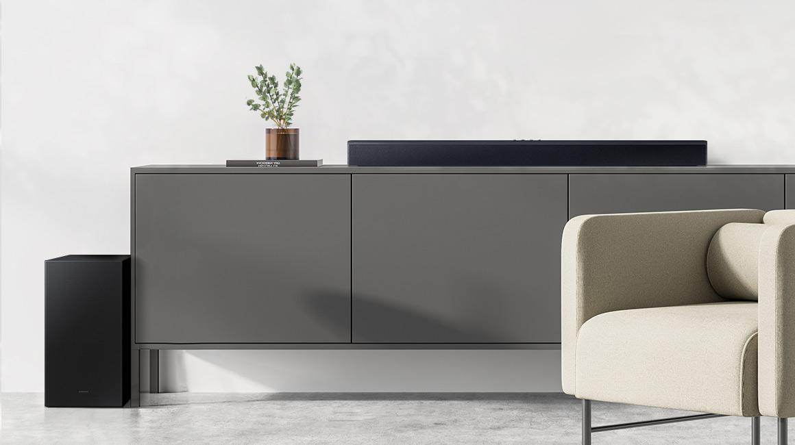 Eine schwarze Soundbar auf einem grauen Schrank. Ein schwarzer Subwoofer steht links. Rechts steht eine Couch.