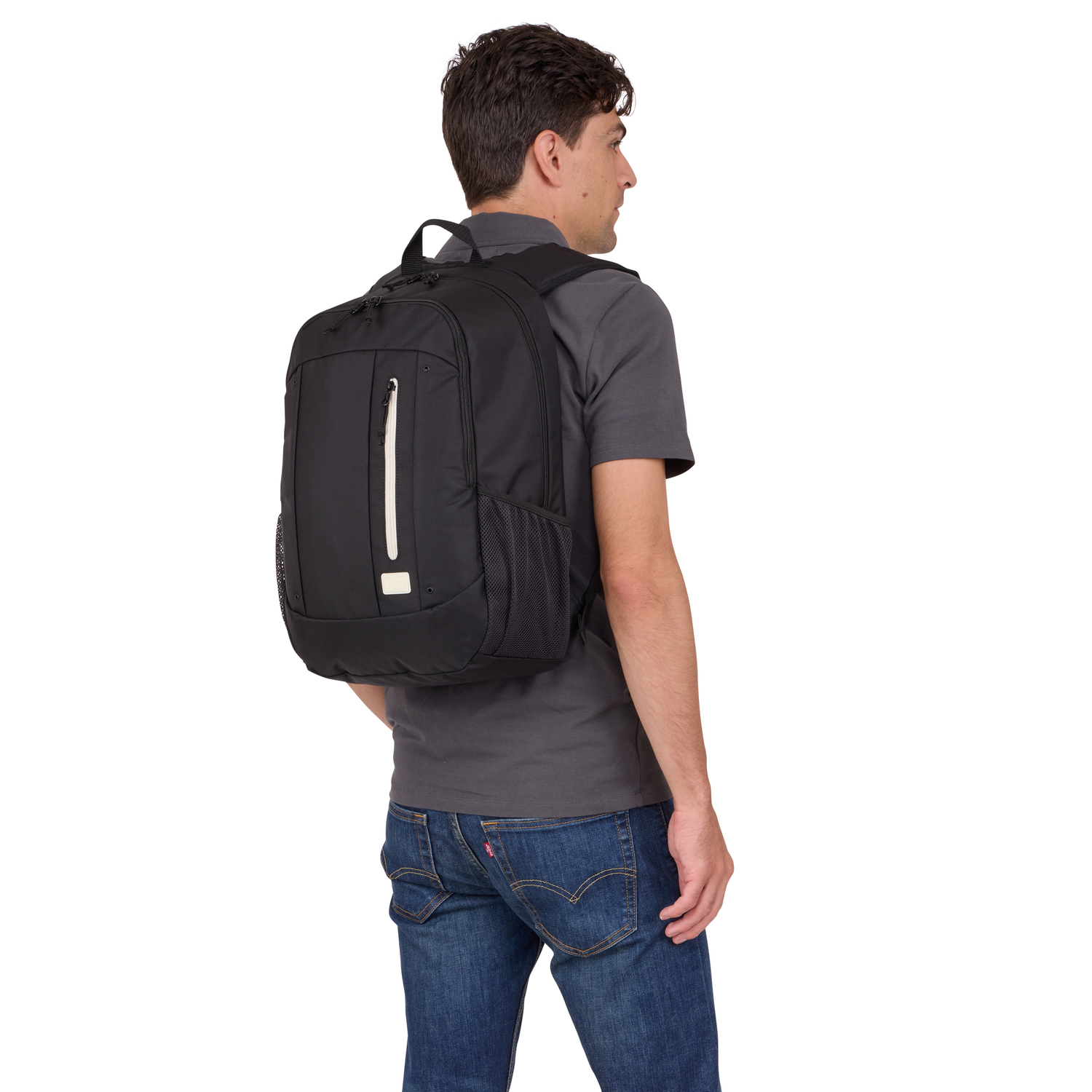 Man with black backpack, wearing grey shirt and jeans, standing against white.
