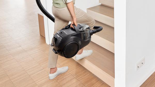 Eine Frau, die einen schwarzen Staubsauger die Holztreppe hochträgt.